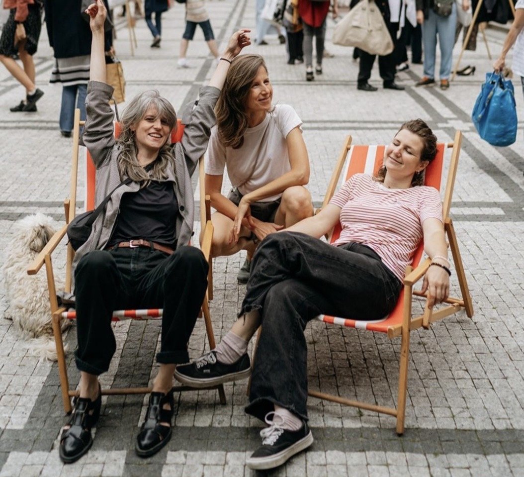 Three women sitting and relaxing in outdoor chairs in a busy street, smiling and enjoying each other's company.