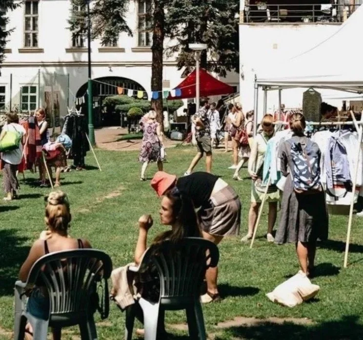 People browsing outdoor market stalls and sitting on chairs on a grassy area, with buildings in the background.