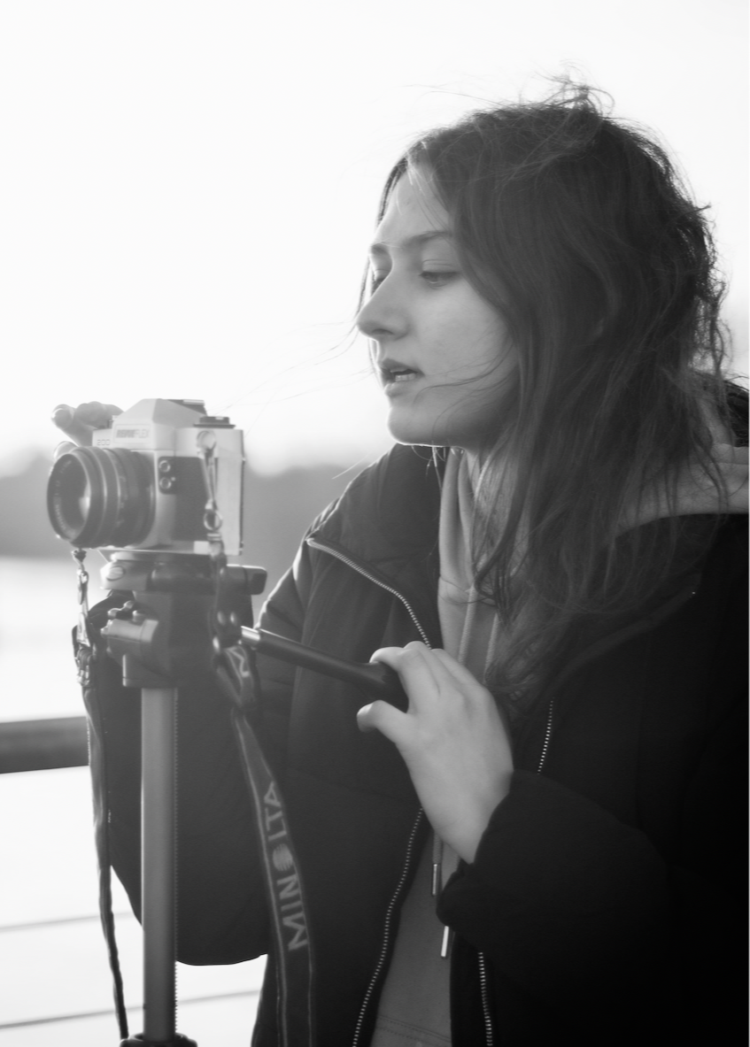 A young woman with long, wavy hair adjusting a camera on a tripod outdoors.