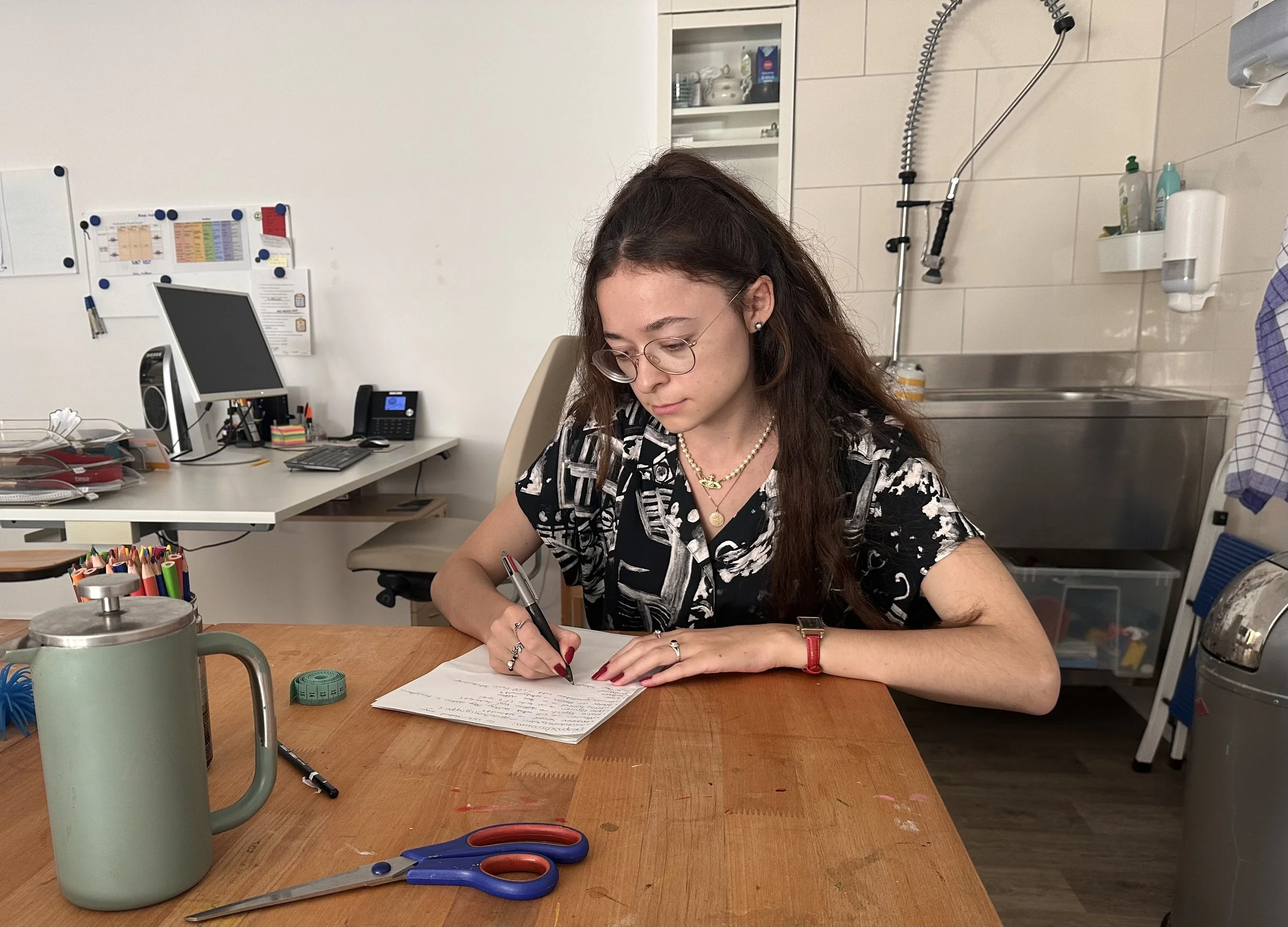 A young woman with long brown hair and glasses is sitting at a wooden table, writing on a sheet of paper with a pen. There are office supplies, including green tape, scissors, and a thermos, on the table. In the background, there is an office desk with a computer, phone, and various papers, as well as a whiteboard on the wall and a kitchen area with a sink, soap, and a towel.