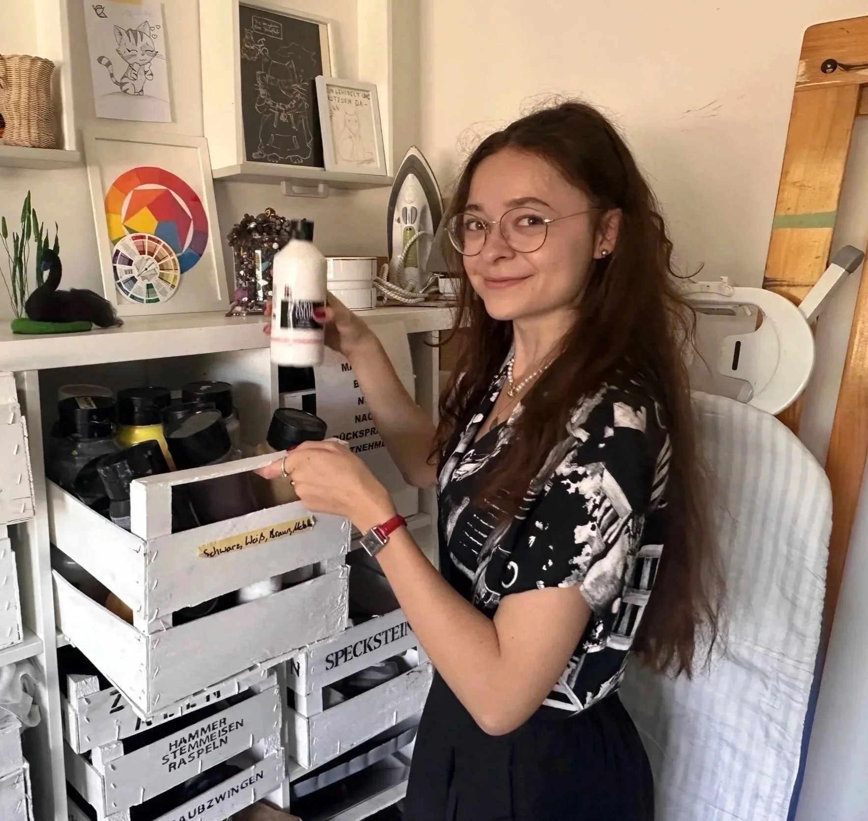 A woman with long curly brown hair, glasses, and a black and white patterned shirt is smiling and holding a bottle of ketchup in a kitchen. She is standing next to a white shelf filled with jars of mustard and ketchup. Behind her, there are framed pictures and illustrations on the wall, including a cute cartoon cat and a color wheel poster. An iron and some papers are also visible on the counter.