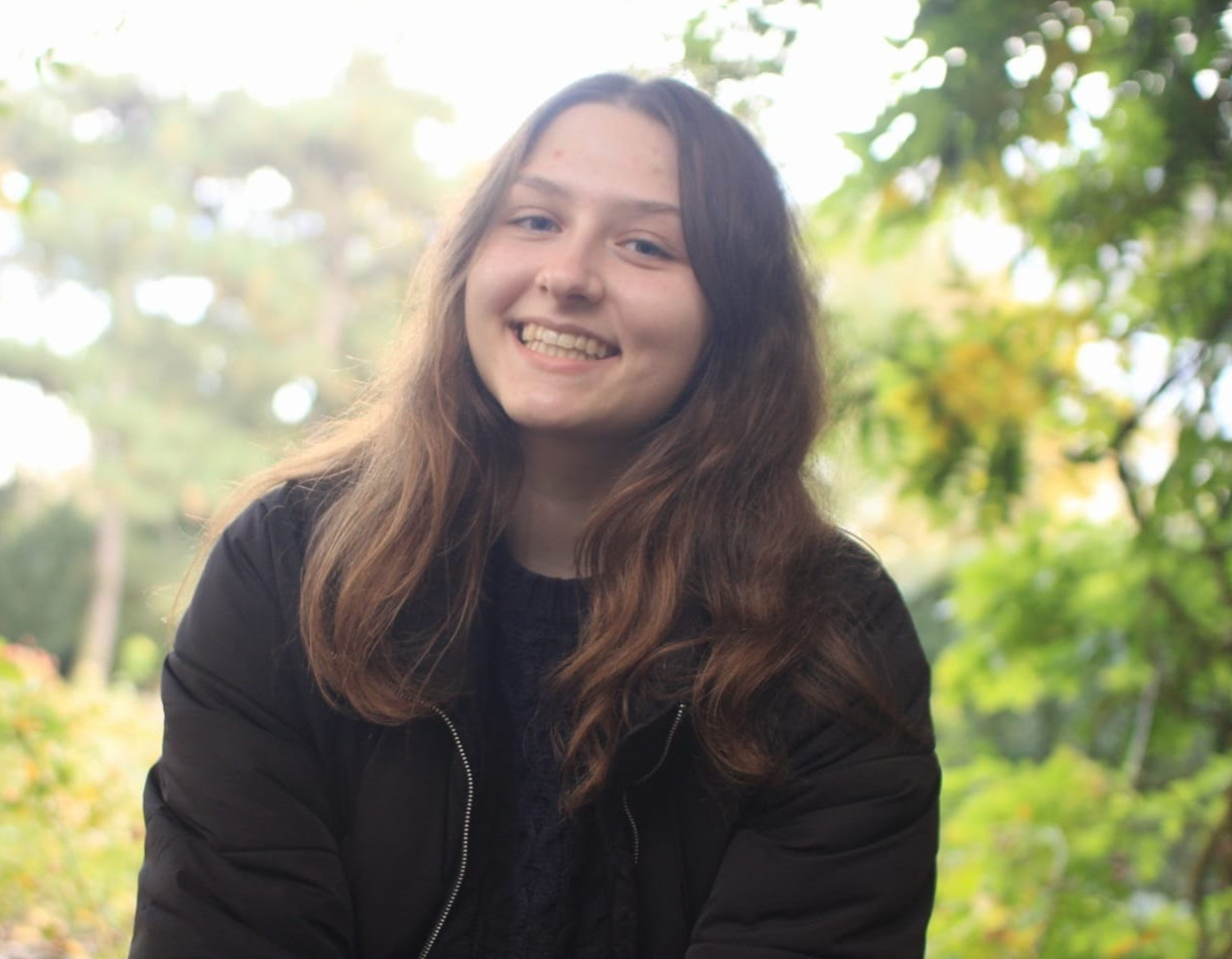 A young woman with long wavy brown hair smiling outdoors in front of green trees and foliage