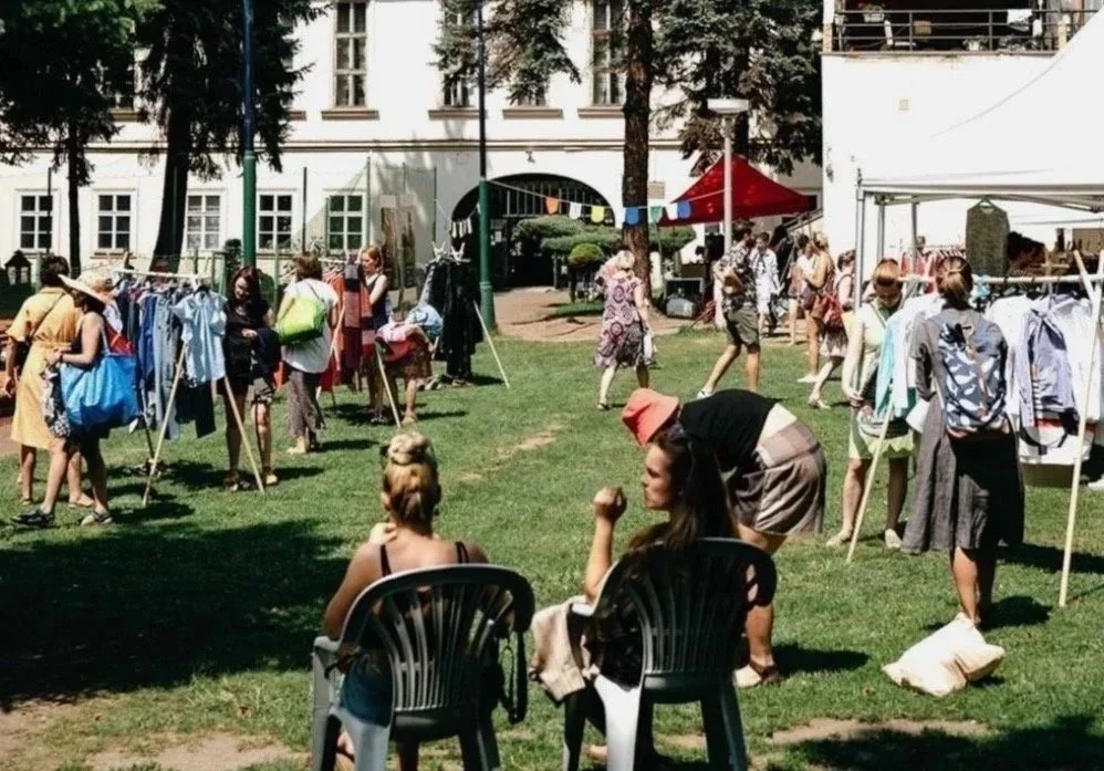Outdoor clothing market with clothing racks, vendors, and shoppers in a park-like setting with trees and a white building in the background.