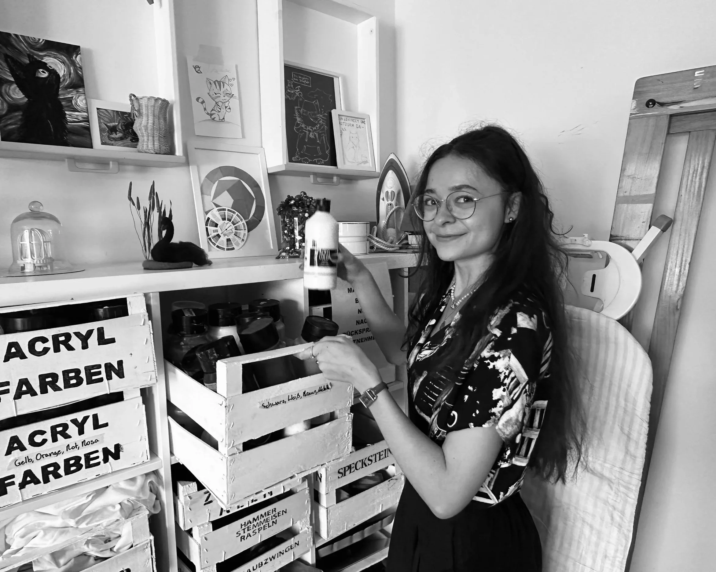 Young woman with glasses and long dark hair smiling at camera in a workshop, holding a can of spray paint. Shelves behind her contain framed artwork of cats and abstract paintings, and boxes labeled with German words like 'FARBEN' and 'SPECKSTEIN'.