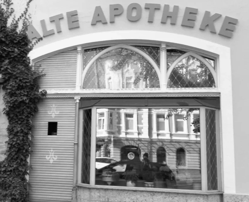 Facade of a building with a large window and decorative arch, with the sign 'ALTE APOTHEKE' above the window, and some plants on the side.
