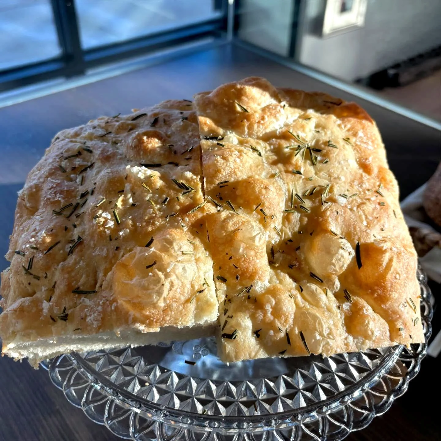 A loaf of baked bread topped with herbs on a glass cake stand.