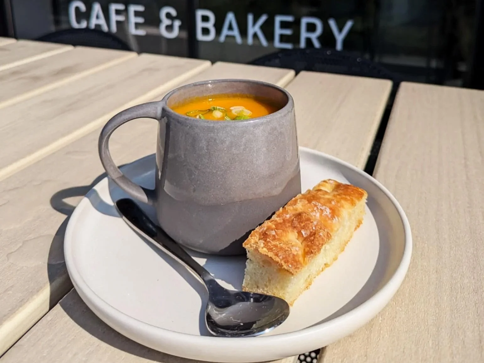 Gray ceramic cup of soup with chopped green onions, a slice of yellow cake on a white plate, and a spoon on a white tray outside at a cafe.