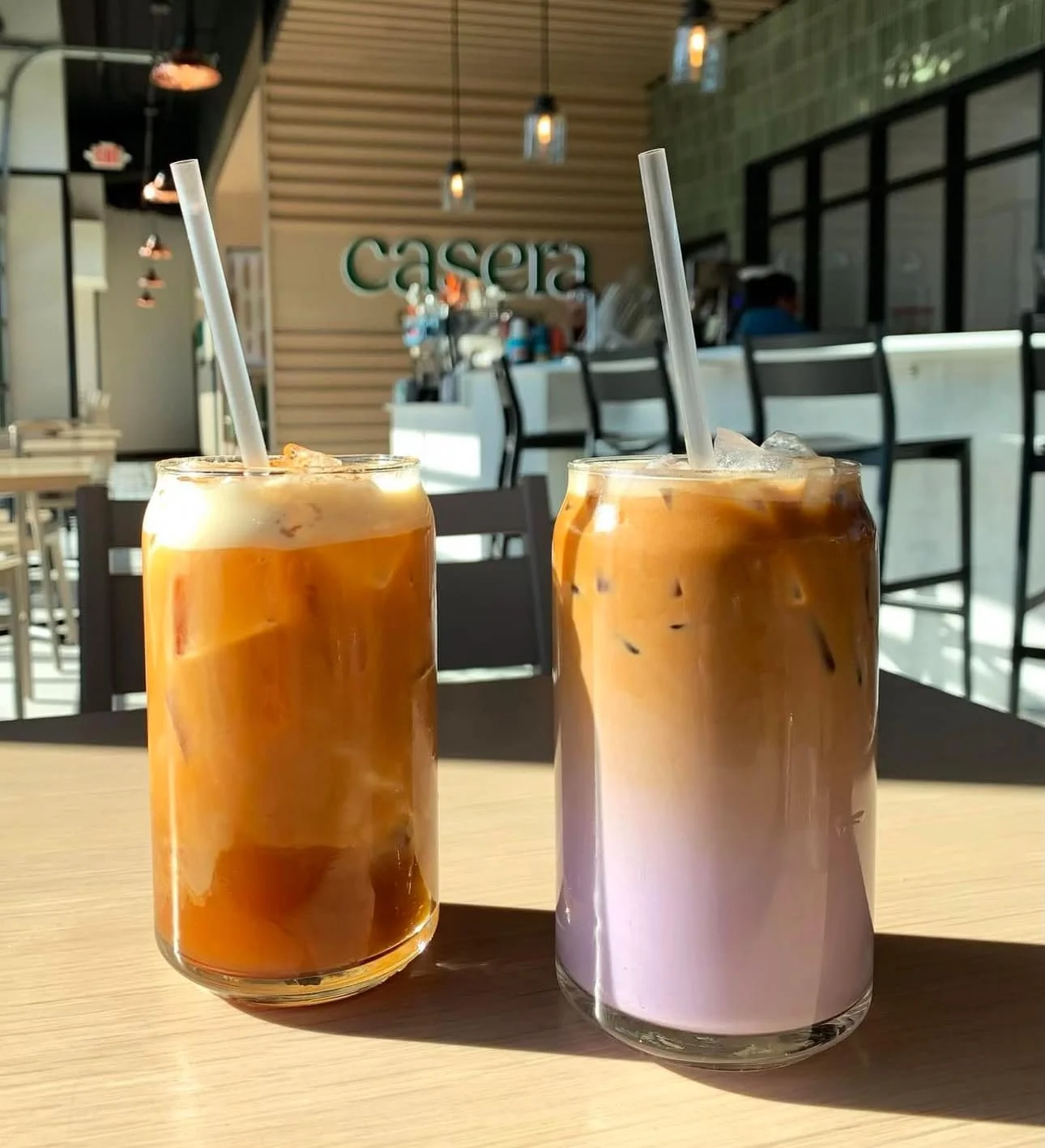Two glasses of iced coffee or iced latte on a wooden table inside a coffee shop, with a blurred background showing the counter and seating area.