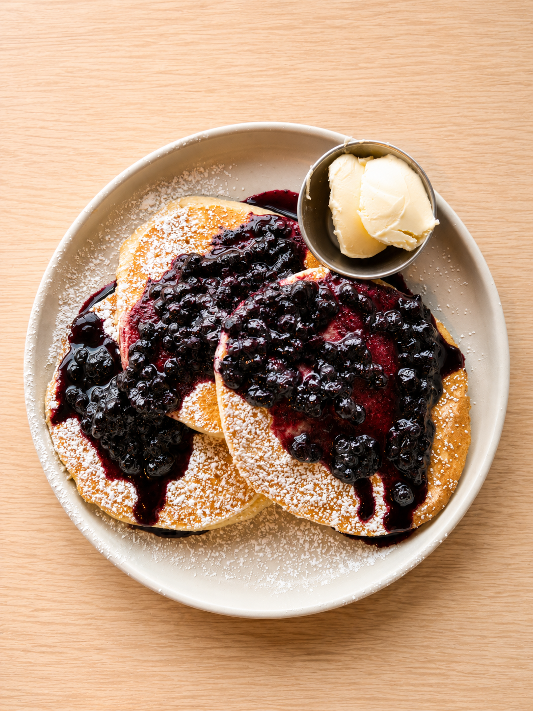 Plate of three pancakes topped with blueberry syrup and powdered sugar, served with a side of butter on a small dish.