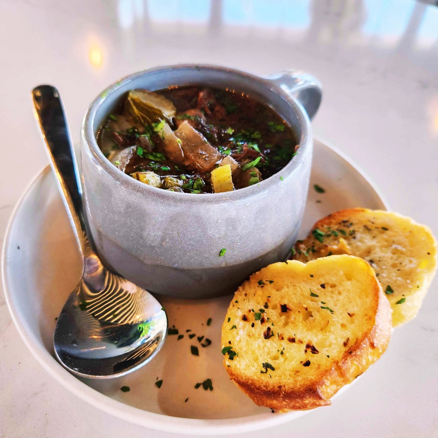 A bowl of beef stew with vegetables and herbs, served with two slices of garlic bread garnished with herbs on a white plate, with a silver spoon placed on the left.