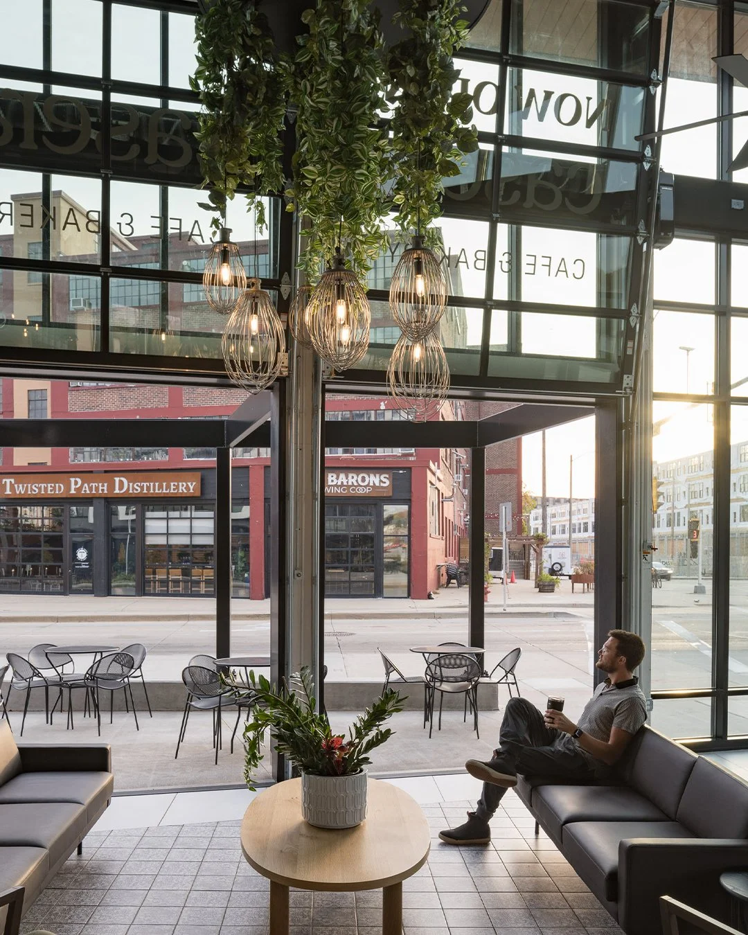 Interior of a modern coffee shop with large glass windows and outdoor seating visible. A man is sitting on a black sofa, holding a cup. Hanging light fixtures and a plant in a white pot are visible, with a city street outside.
