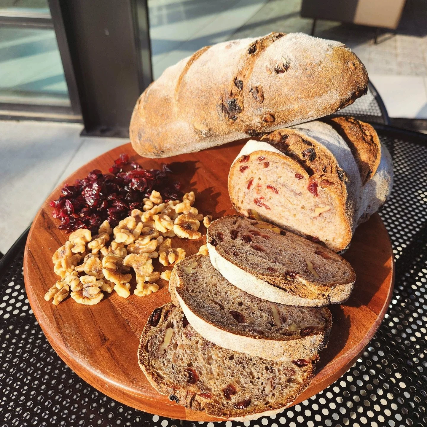 Sliced fruitcake bread with dried fruit and nuts on a round wooden plate, accompanied by dried cranberries and walnuts, outdoors on a sunny day.