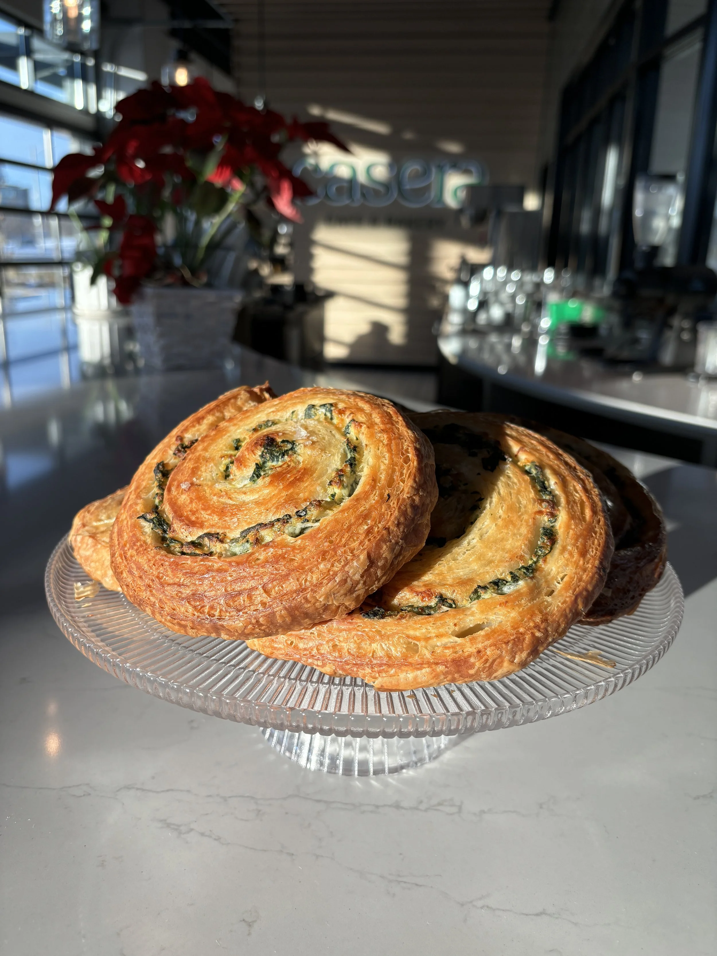 Close-up of three spinach and cheese filled puff pastries on a glass platter at an airport cafe with sunlight streaming through windows.
