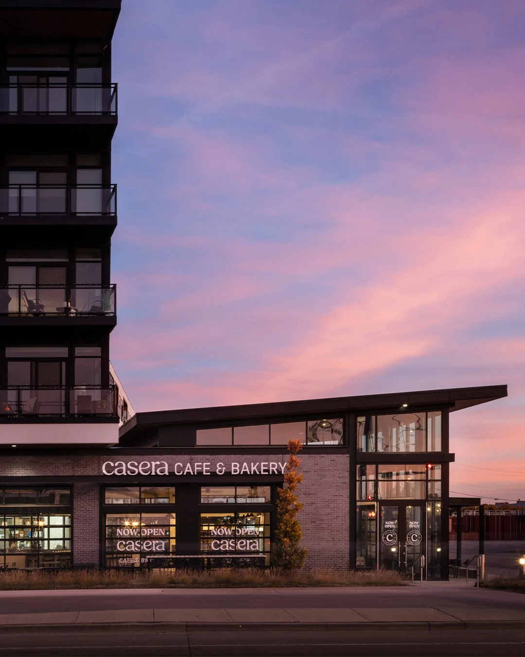 Exterior view of a modern building with a cafe and bakery named 'Casera' during sunset, with a pastel sky in the background.