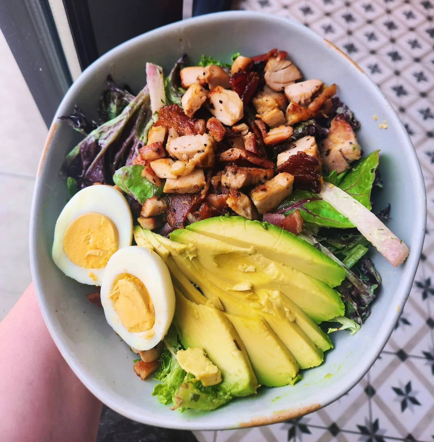 Bowl of salad with mixed greens, sliced avocado, hard-boiled egg halves, diced grilled chicken, and bacon strips, on a decorative black and white tiled floor.