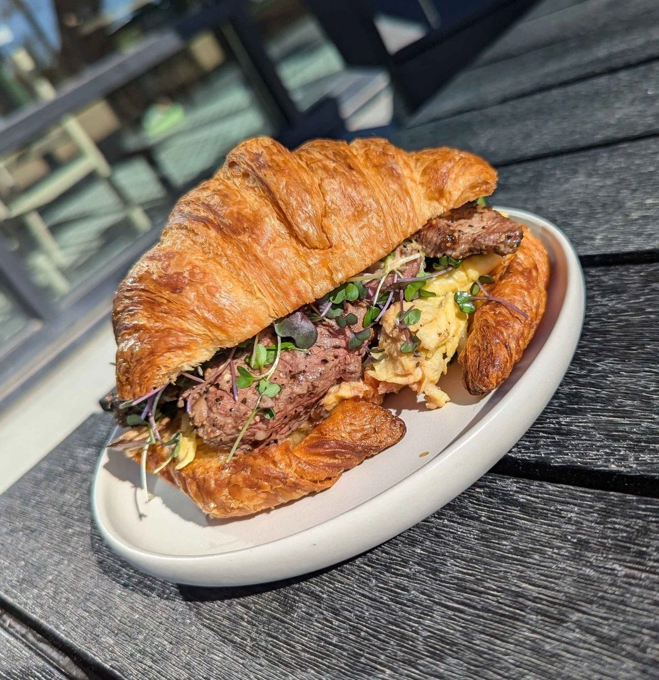Croissant sandwich with beef patty, scrambled eggs, microgreens, and other ingredients on white plate outside on a wooden table.