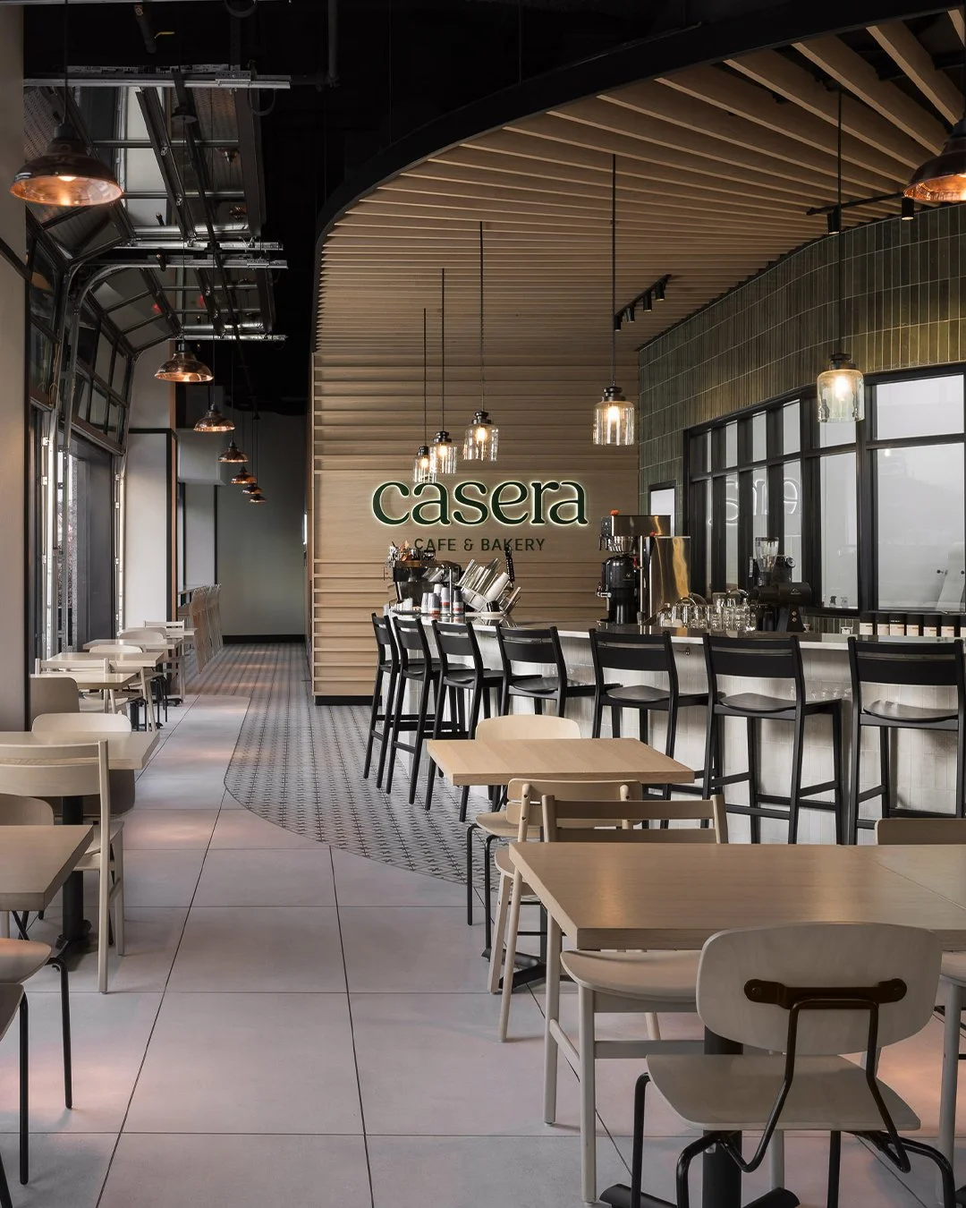 Interior of a modern cafe named 'Casera' with a barista counter, bar stools, and small tables with chairs, windows on the right, and decorative lighting.