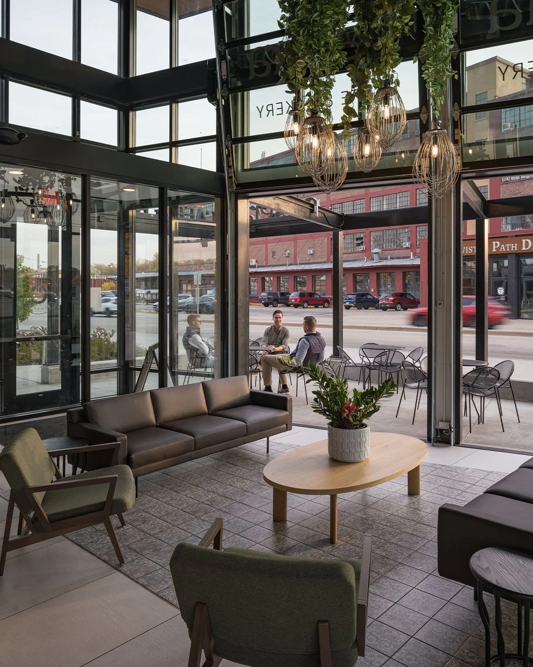 Interior view of a modern coffee shop or cafe with large glass windows and doors, a brown leather sofa, green armchairs, a wooden coffee table with a potted plant, and contemporary hanging light fixtures. Outside, three men sit at outdoor tables on the sidewalk, with a street and brick buildings visible in the background.