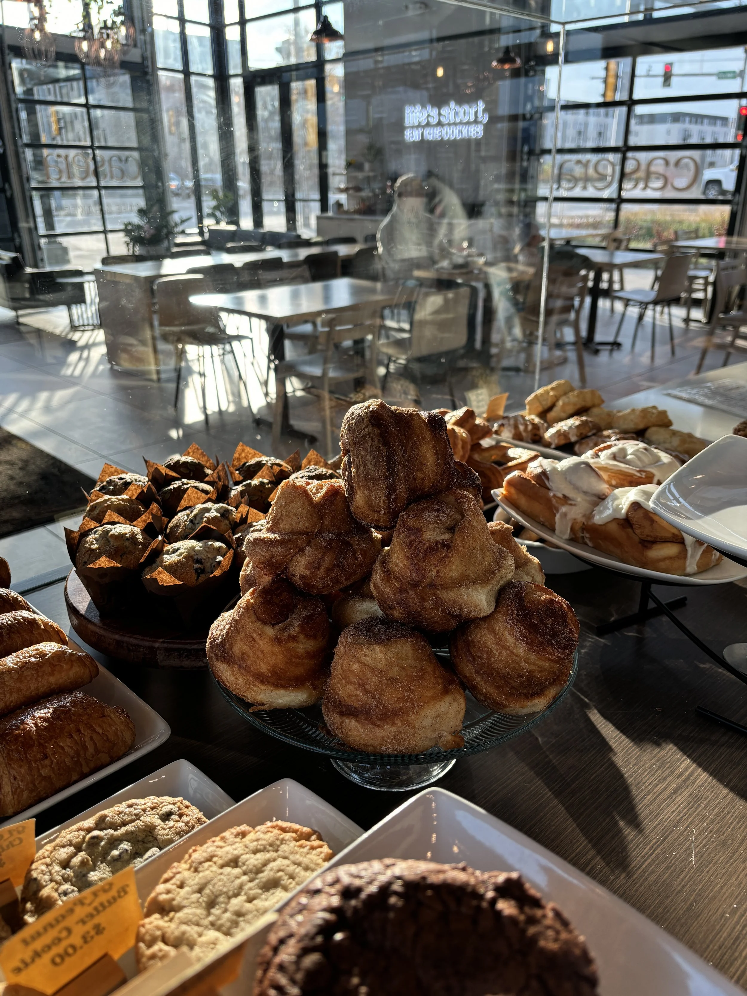 Display of assorted desserts including donuts, cookies, and pastries inside a bakery or cafe, with sunlight streaming through large windows.