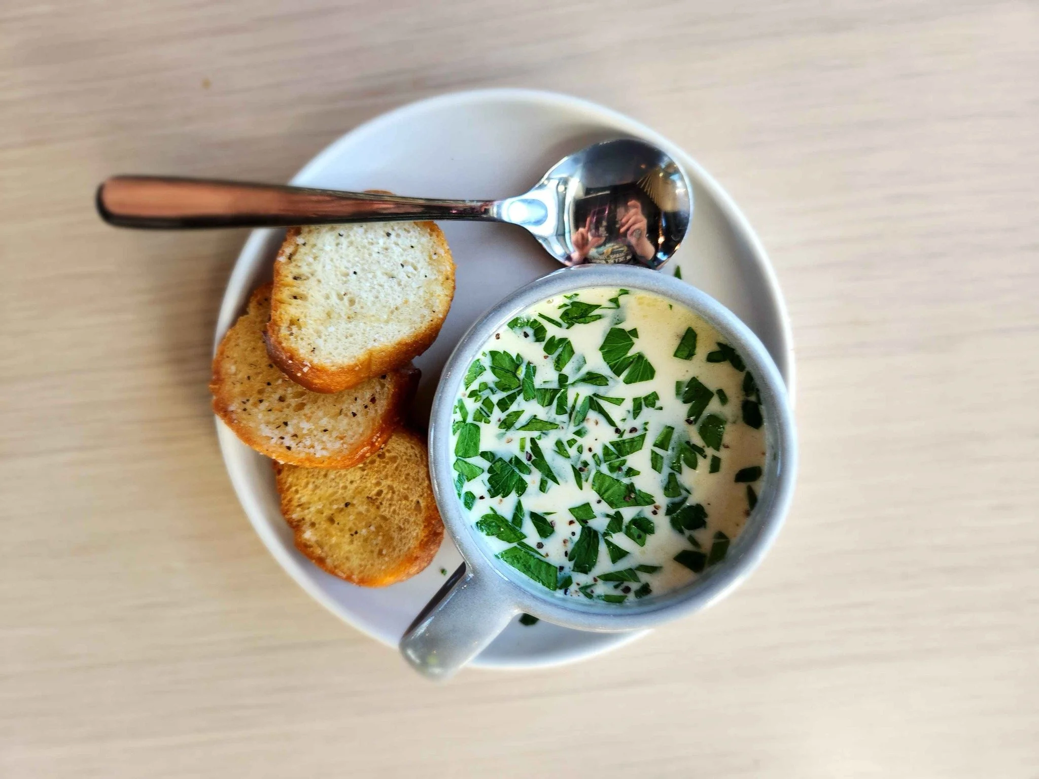 A white bowl of creamy soup garnished with chopped green herbs, placed on a light wooden table. Next to the bowl, there are two slices of toasted bread and a metal spoon resting on the bowl.