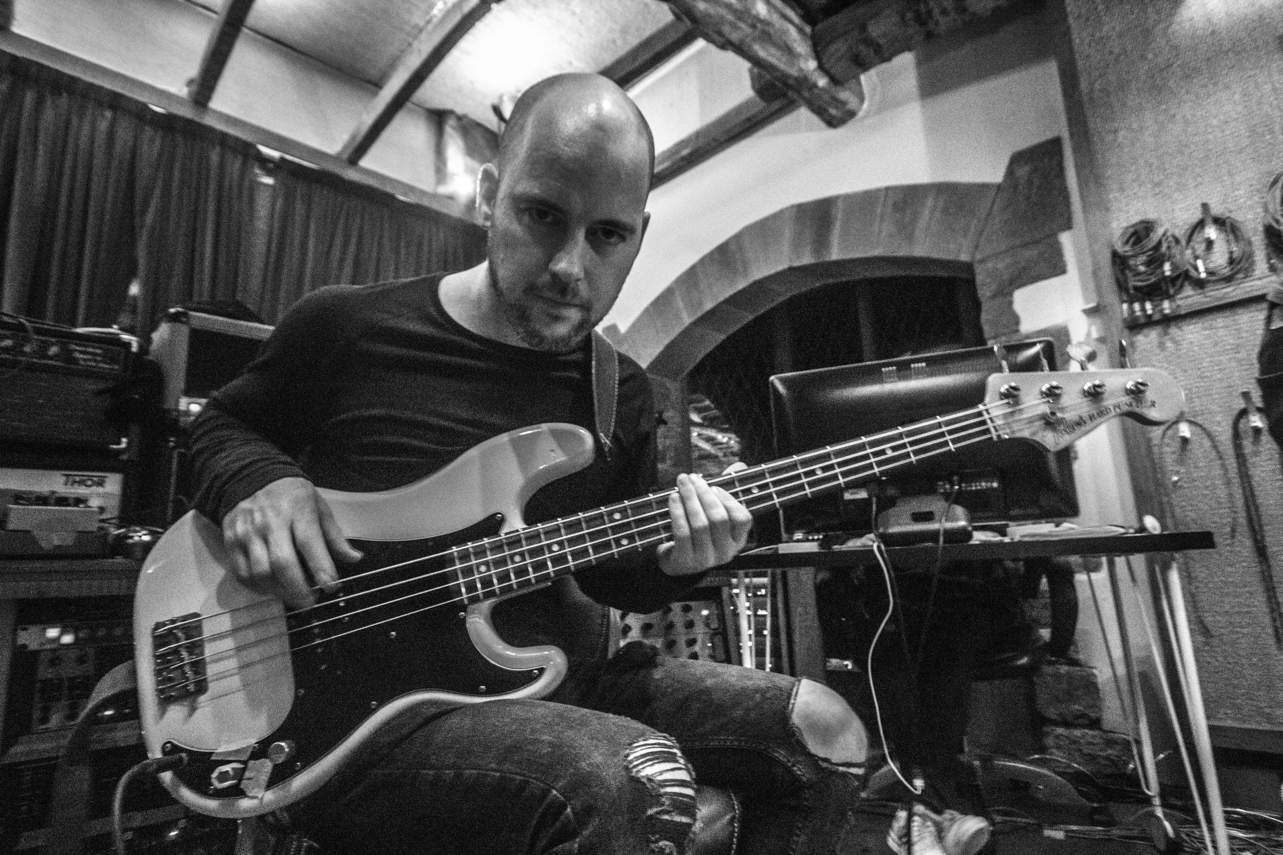 A man with a shaved head playing an electric bass guitar in a music studio, surrounded by musical equipment and instruments.