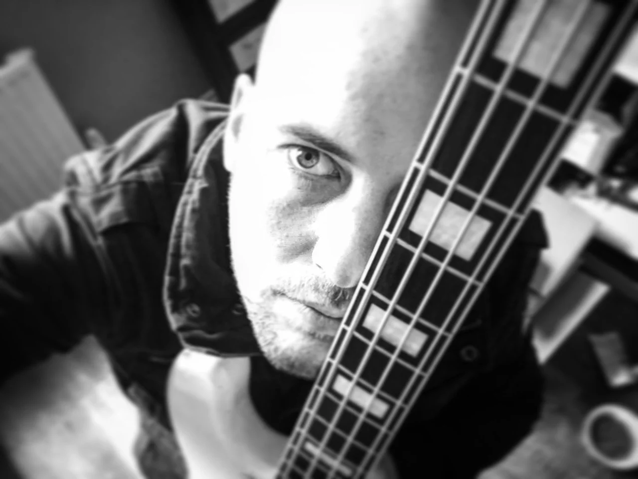Black and white close-up photo of a man with light-colored eyes and light facial hair, holding a guitar vertically in front of his face, partially obscuring his right eye, in an indoor setting.