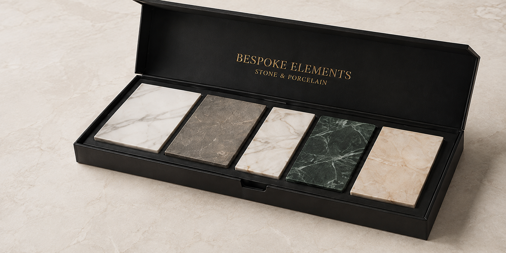 A black box with gold lettering that says 'Bespoke Elements Stone & Porcelain' contains five rectangular stone and porcelain tiles in various colors and patterns, arranged neatly inside.