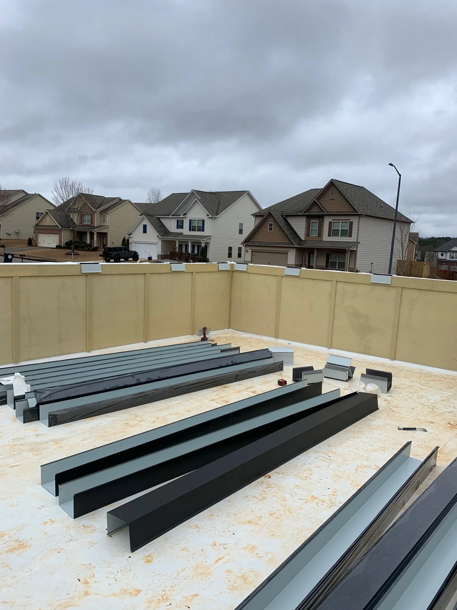 View from a construction site with metal roofing panels and tools, overlooking a row of residential houses under cloudy sky.