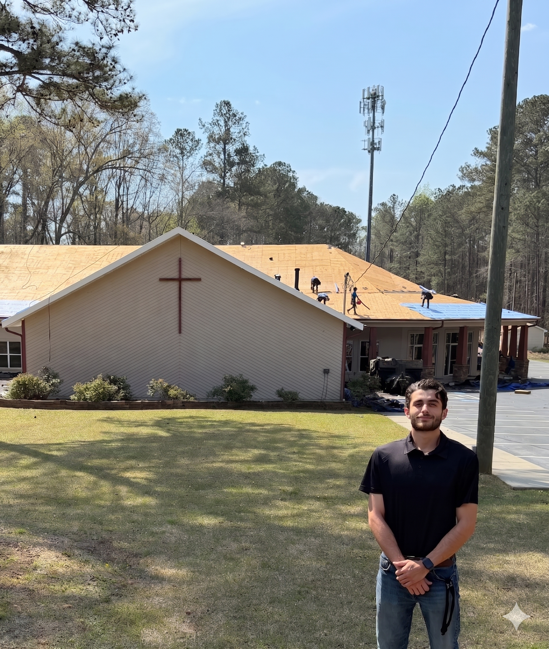 A young man in a black shirt and jeans standing outdoors in front of a church with a cross on its roof. The church is under construction, with workers on the roof and a new metal roof being installed. Trees and a cell tower are visible in the background.