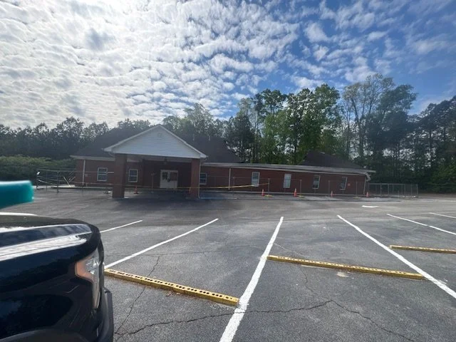 Empty parking lot in front of a single-story brick building with a white front porch, surrounded by green trees under a partly cloudy sky.