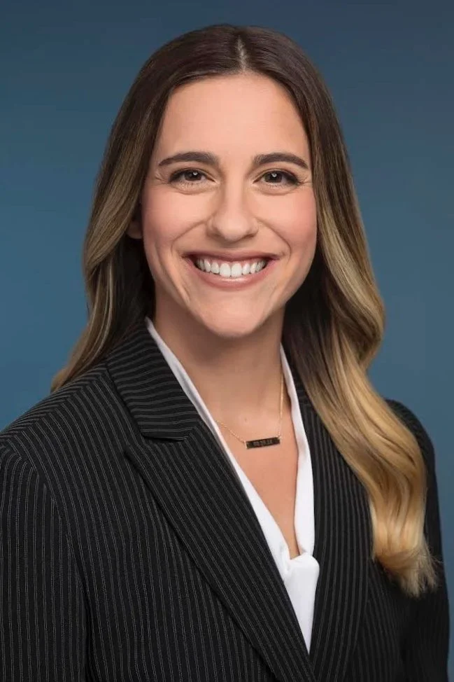 Professional woman with long wavy hair, smiling, wearing a black pinstripe suit and white blouse, against a plain blue background.