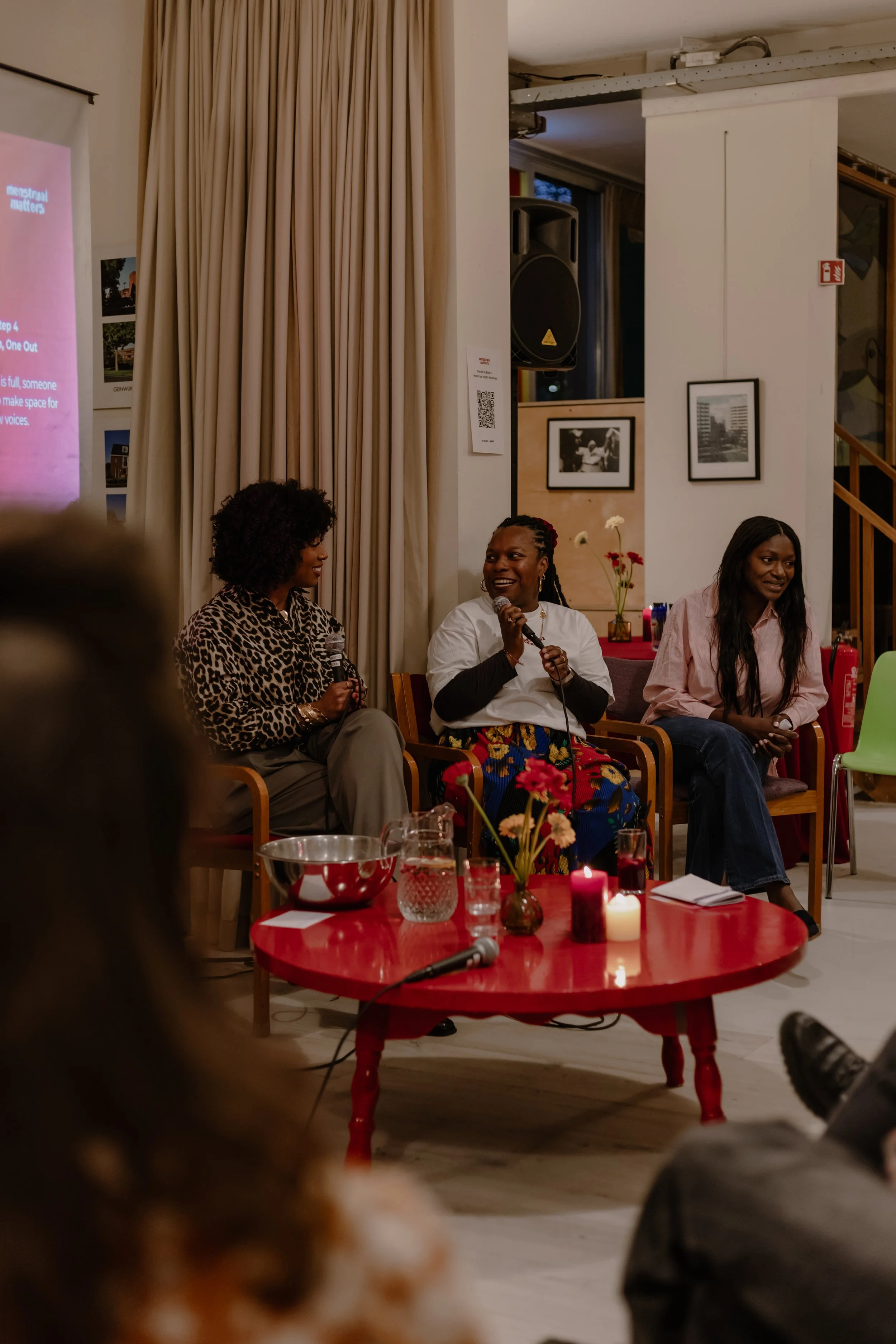 Three women sitting on chairs, one holding a microphone, engaging in a discussion or presentation. There are framed pictures on the wall and a red table with candles and flowers in the foreground.