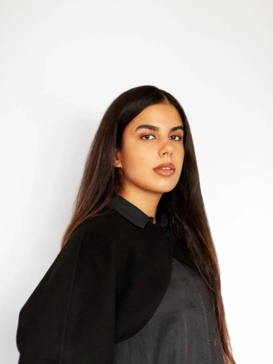 Portrait of a woman with long dark hair wearing a black top, standing against a plain white background.