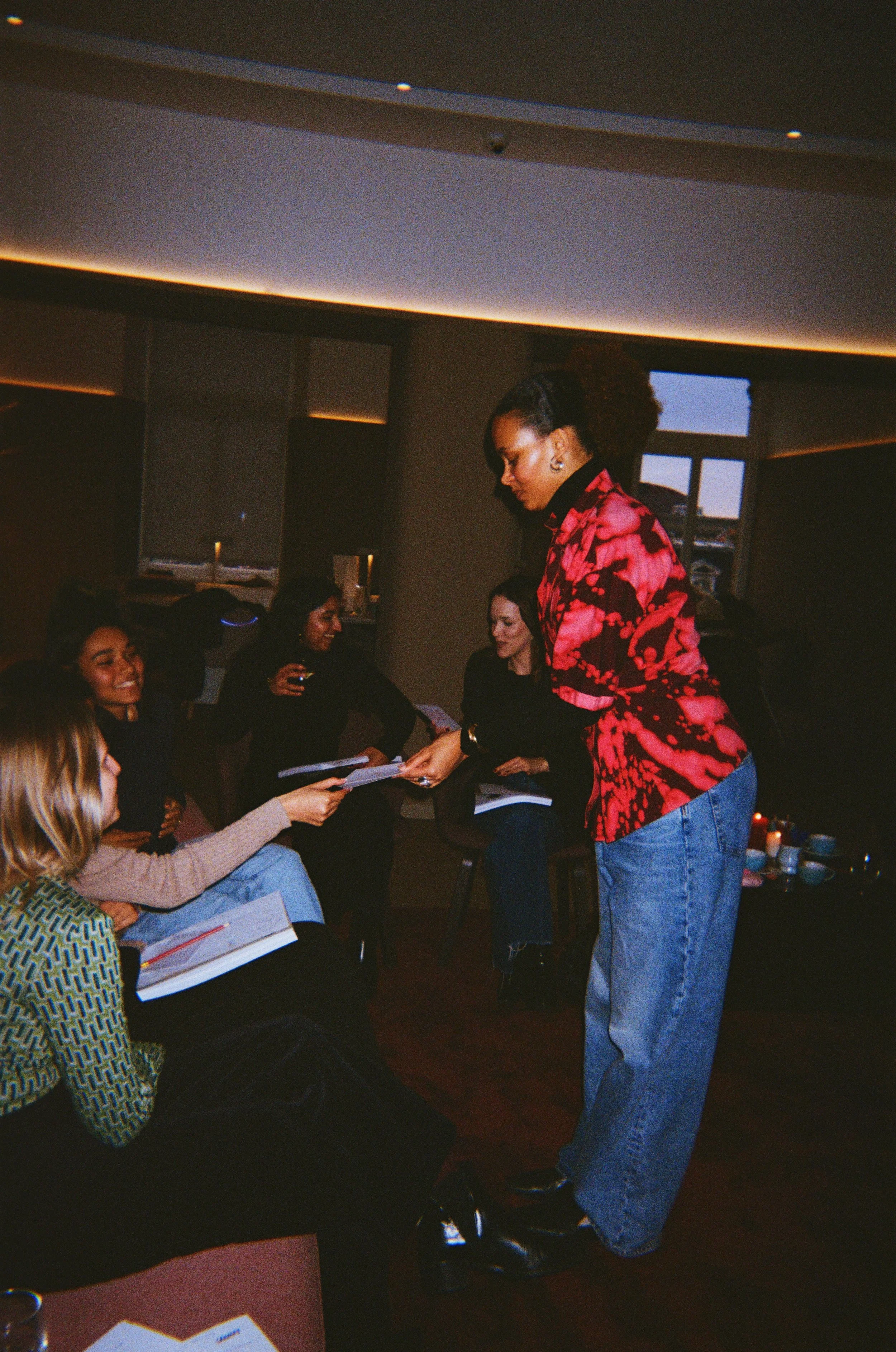 A woman in a red and black patterned shirt is giving a certificate to another woman in a beige sweater at an indoor event, with multiple people seated and a window in the background.