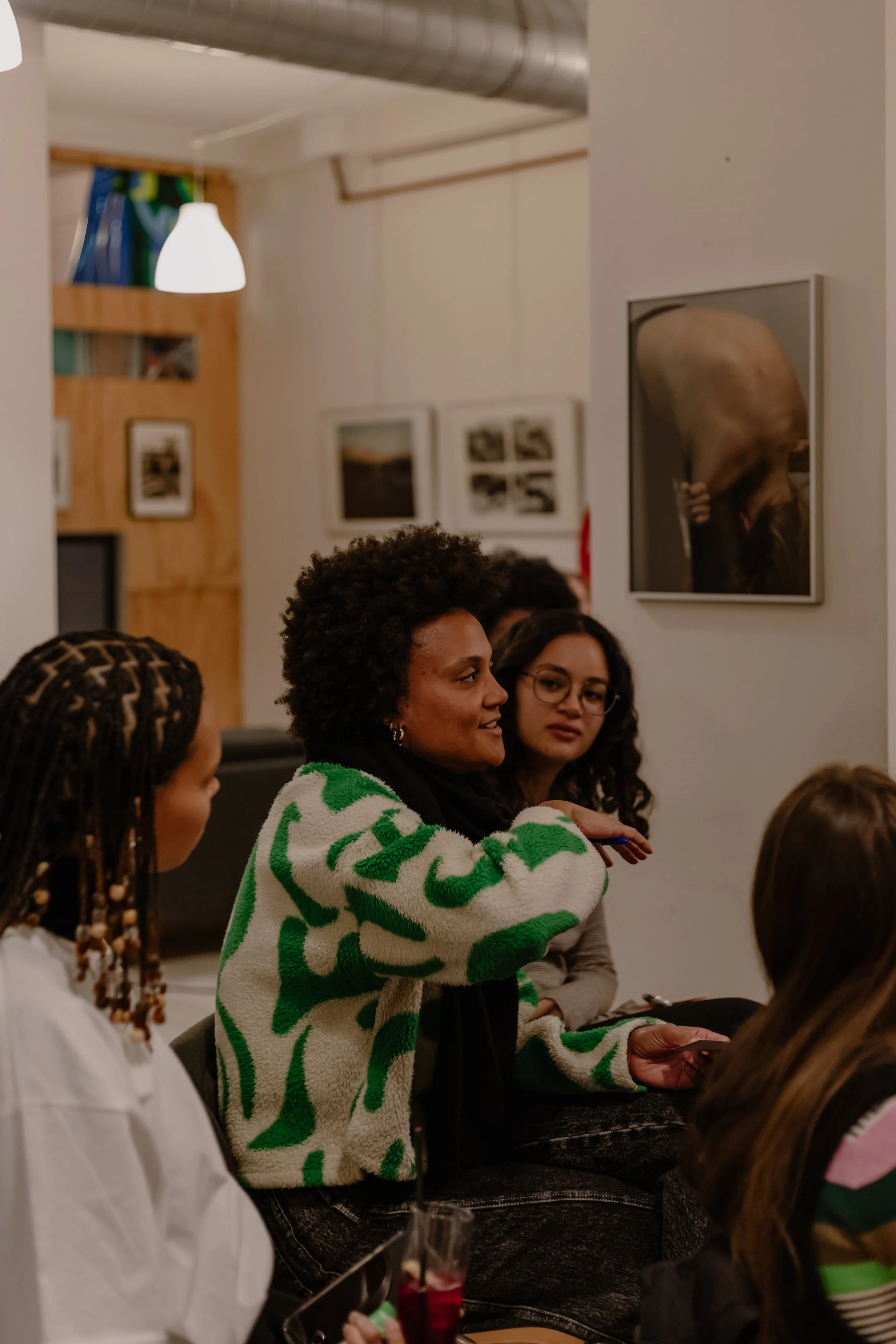 A group of diverse women, including a woman with curly hair in a green and white patterned sweater, gathered in a room with framed photographs on the wall, engaged in conversation.