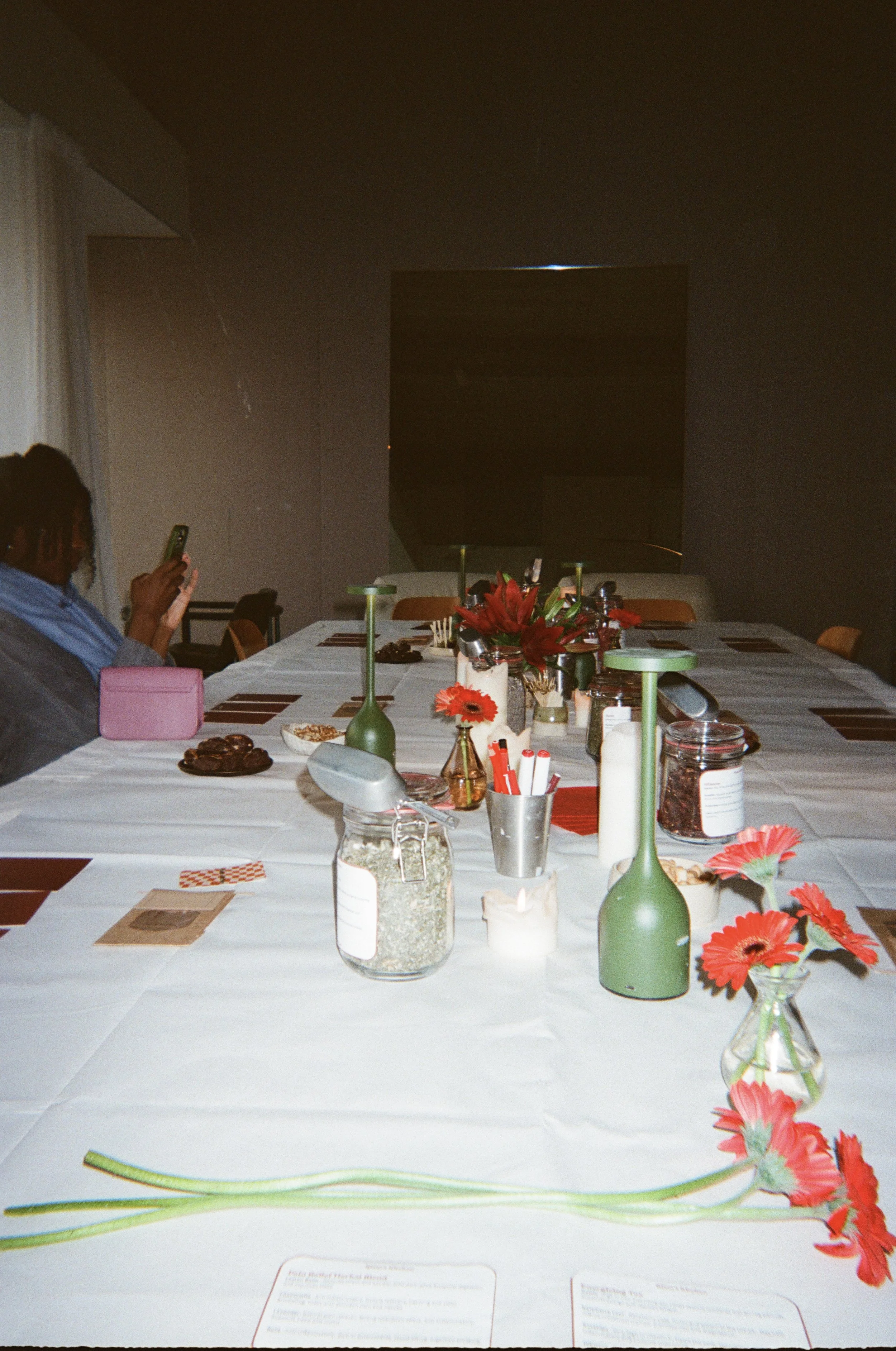 A dining table decorated with red flowers, candles, and various small items, with a woman sitting on the left taking a photo with her phone, in a room with a plain wall and a large television in the background.