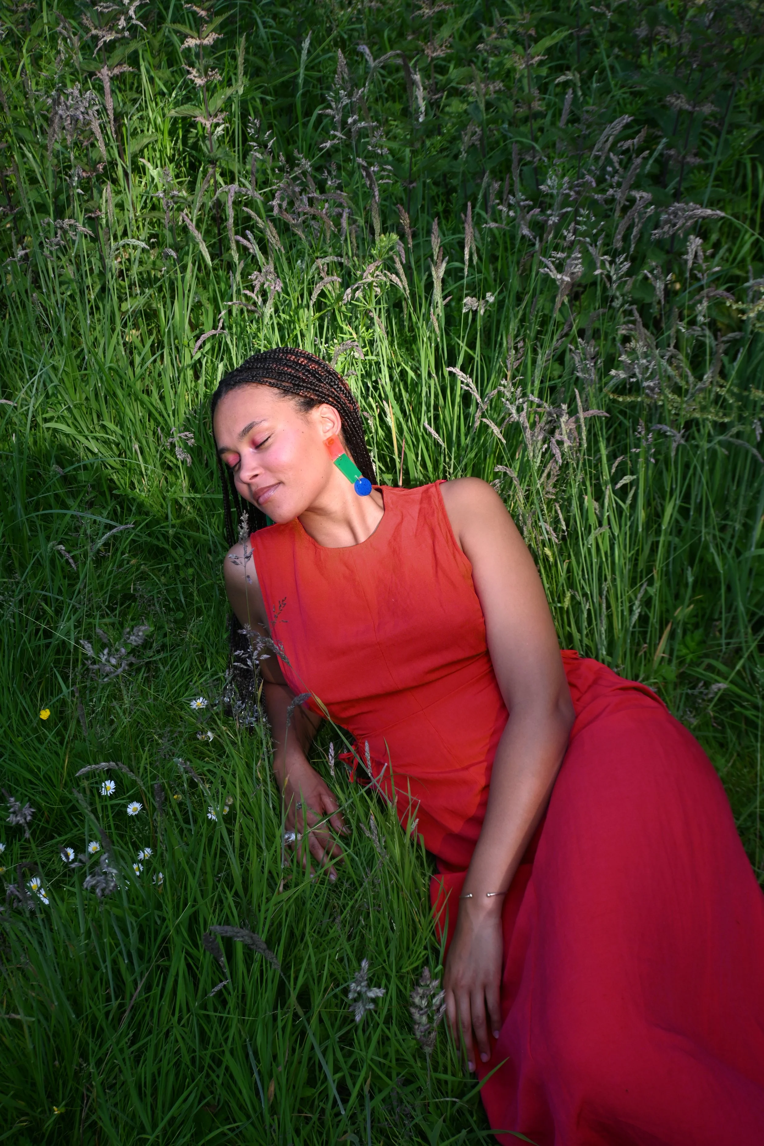 A photo of CRAMPY founder Alizée, wearing a red dress, is lying in a grassy field with closed eyes.