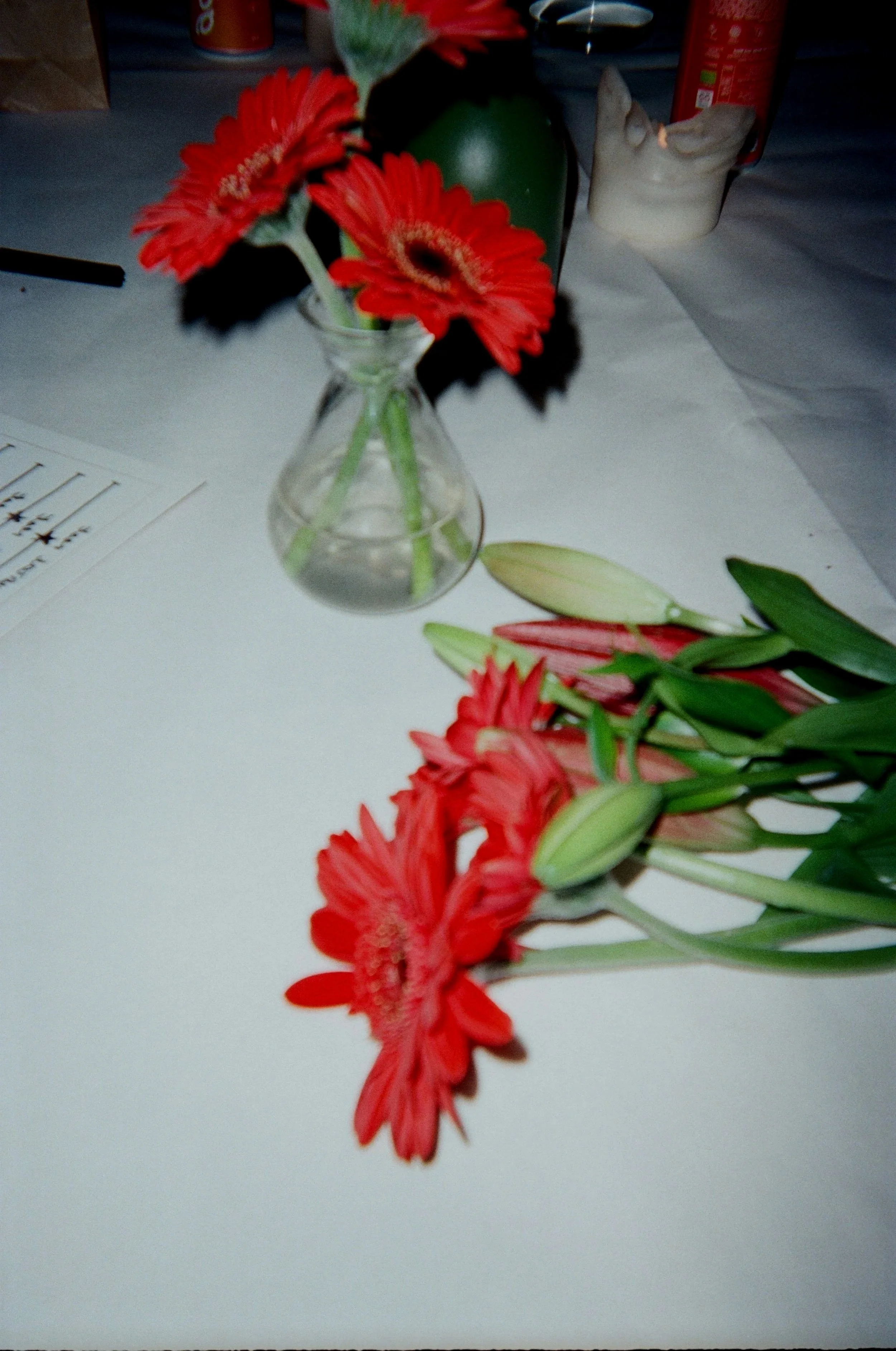Red gerbera daisies arranged in a small glass vase with colored flowers and green leaves, placed on a white surface with various objects and papers around.