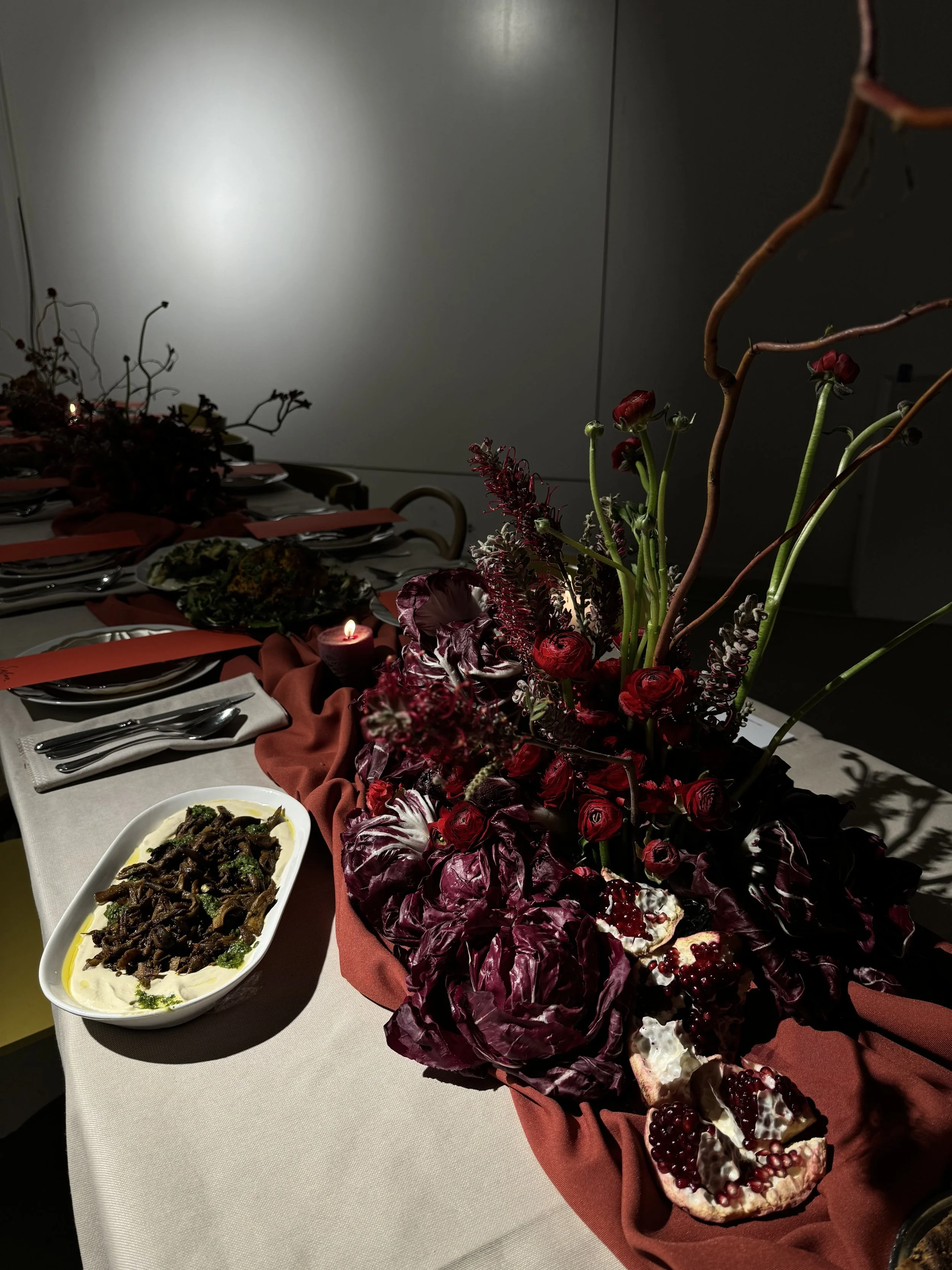 A table with a floral centerpiece and various dishes, including a bowl of salad with greens and a dark topping, set with napkins, silverware, and a red cloth runner in a dimly lit room.