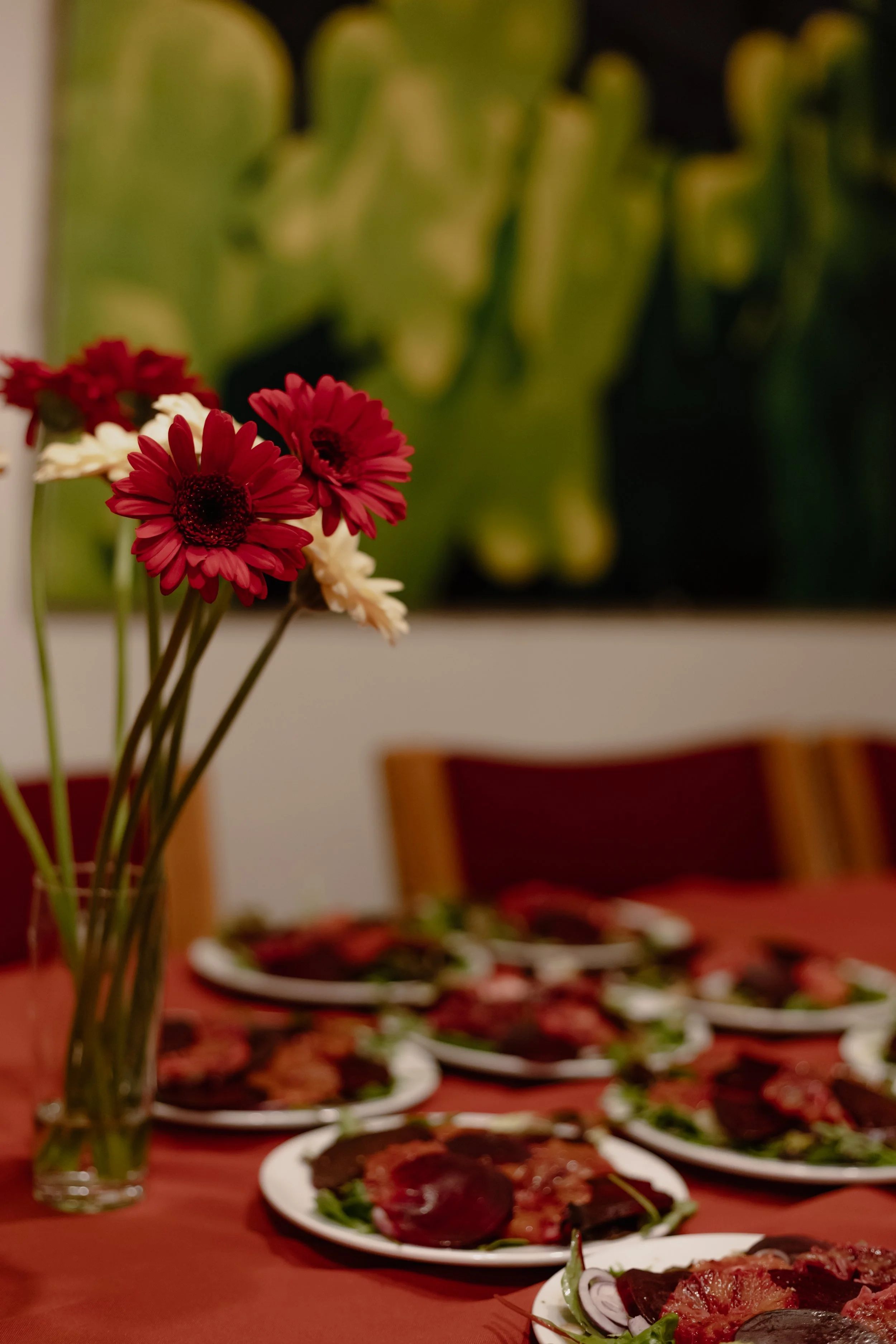 A table set with plates of sliced meats, vegetables, and a vase of red and white flowers, with a blurred background of chairs and a wall with decorative artwork.