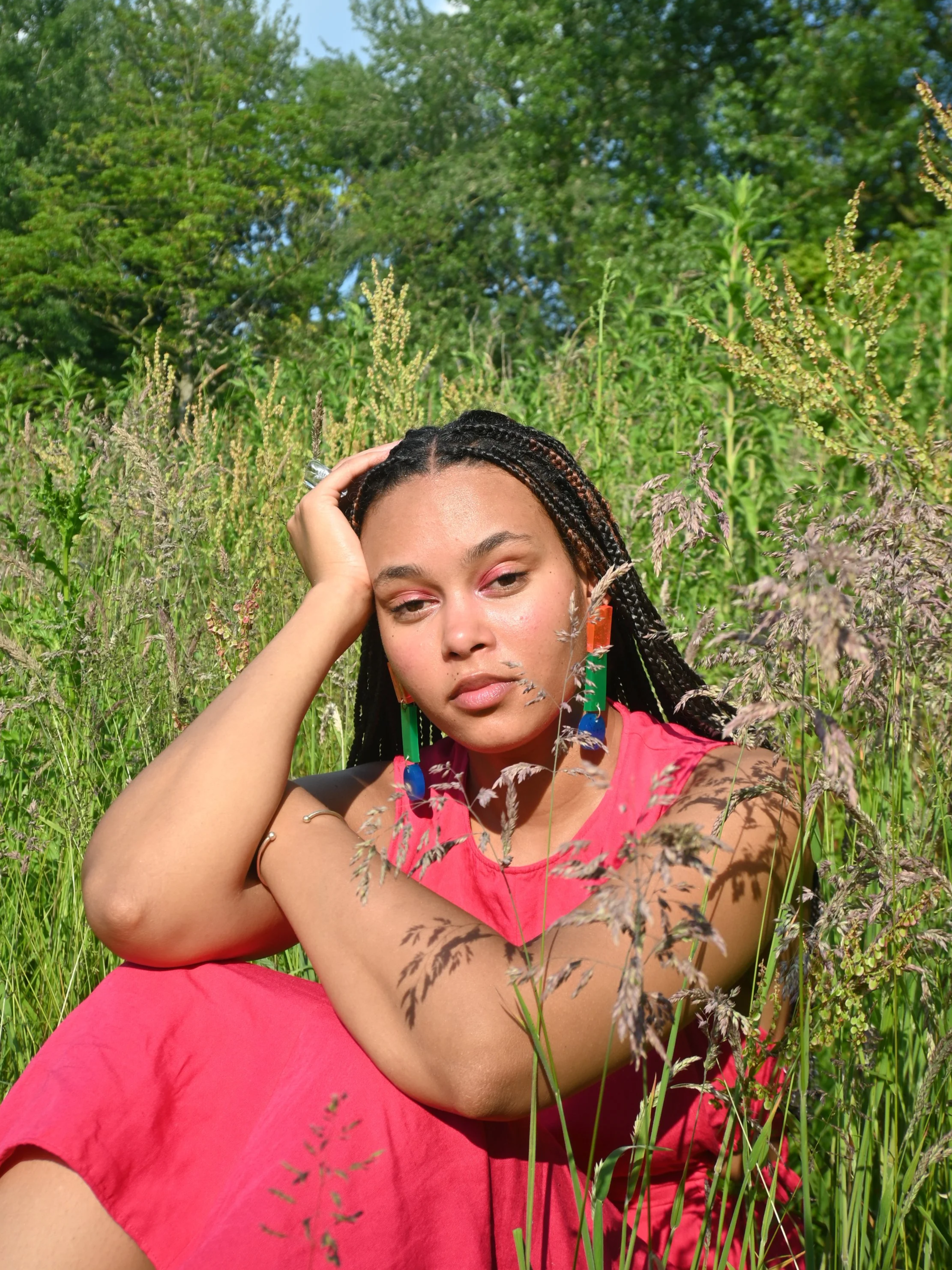 A photo of Alizée Bollen, founder of CRAMPY with braided hair and colorful earrings sitting in tall grass in a green outdoor setting, wearing a pink dress and resting her head on her hand.