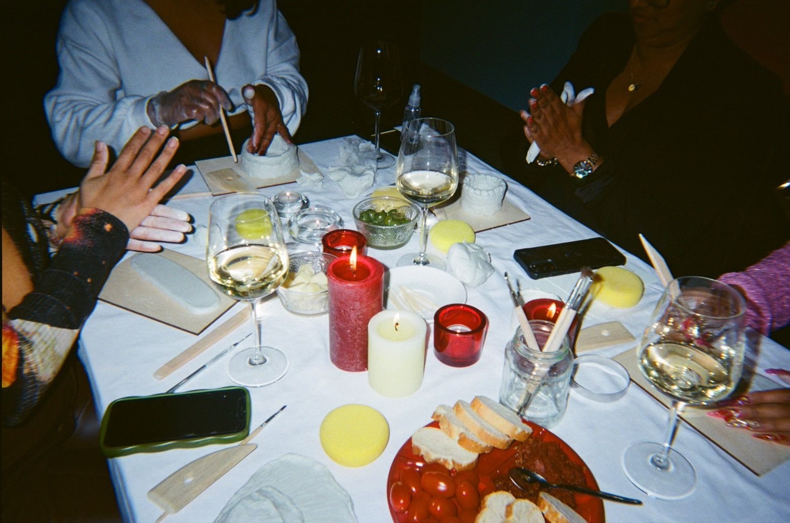 People gathered around a table with candles, wine glasses, cheese, bread, and snacks, celebrating in a dimly lit setting.
