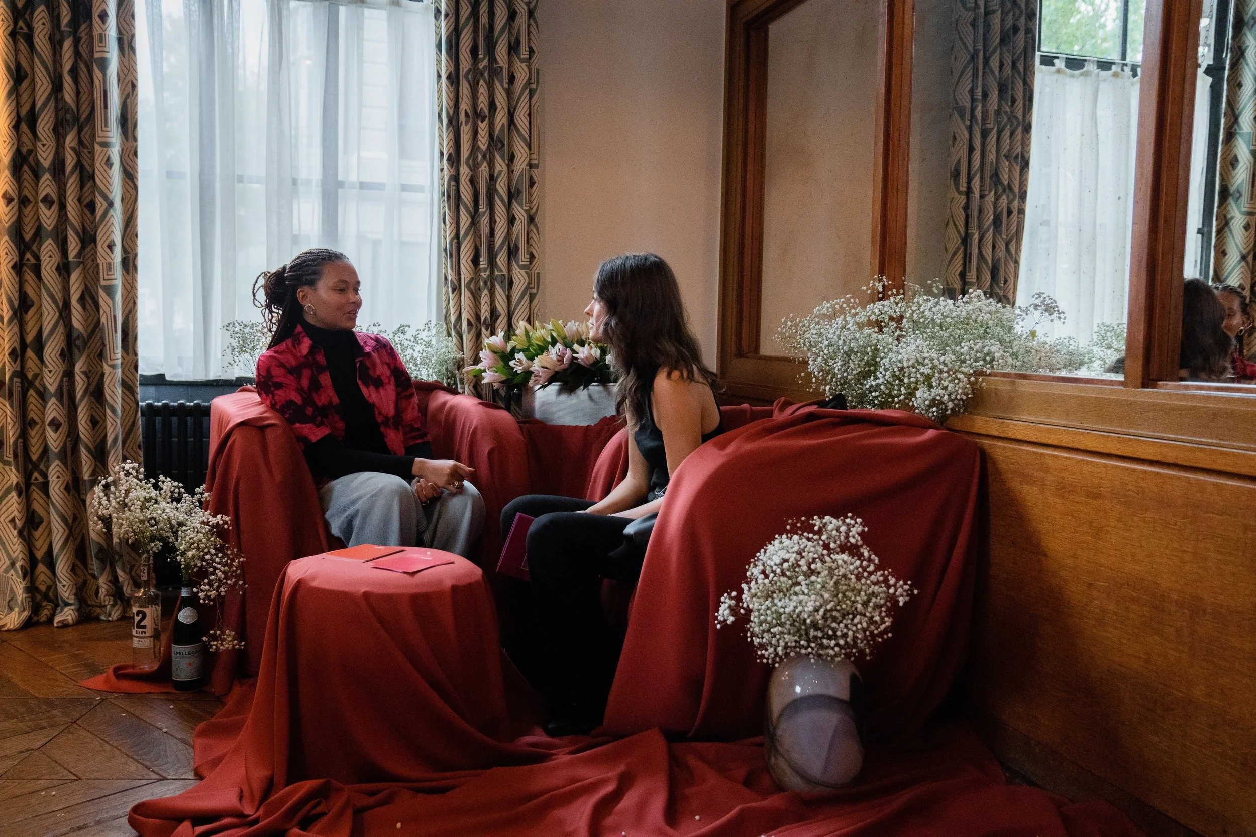 Two women sitting on red-covered chairs in a cozy room, engaged in conversation with floral decorations around them, and a mirror reflecting their image.