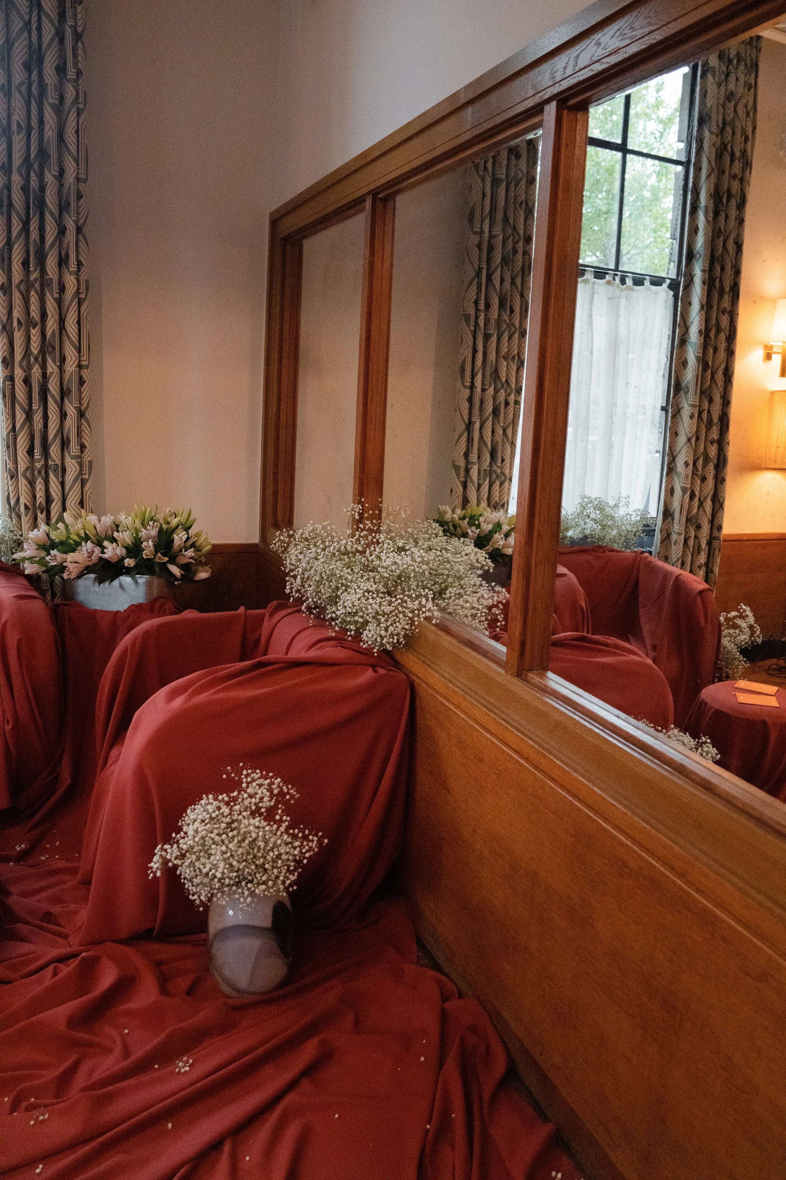 A floral arrangement with white baby's breath and pink flowers placed on red draped furniture, viewed through a wooden frame in a room with patterned curtains and a window showing green trees outside.