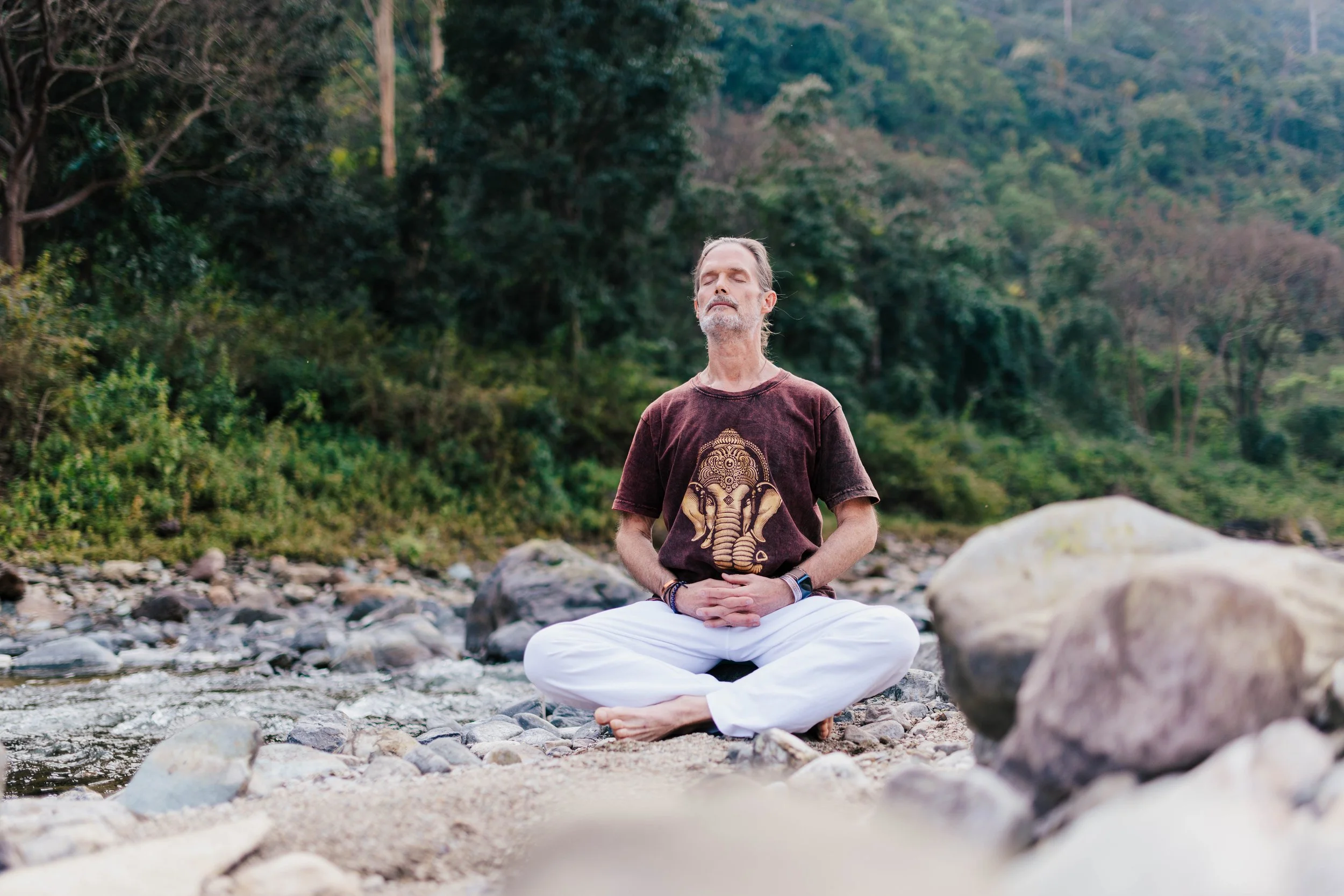 Bill sitting cross-legged on rocks by a river, meditating with eyes closed, wearing a brown T-shirt with a golden elephant design, surrounded by green trees and mountains.