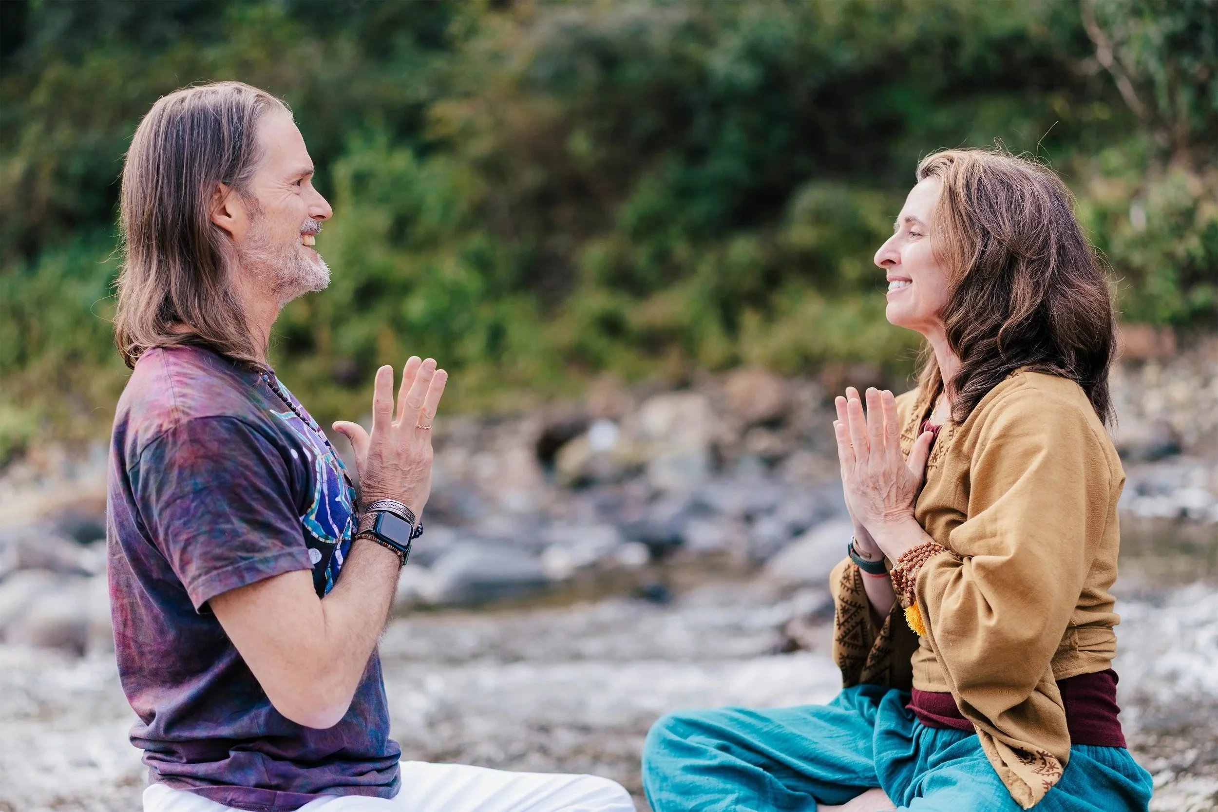 Bill and Lexie face each other, sitting on a beach, with their palms together and smiling.