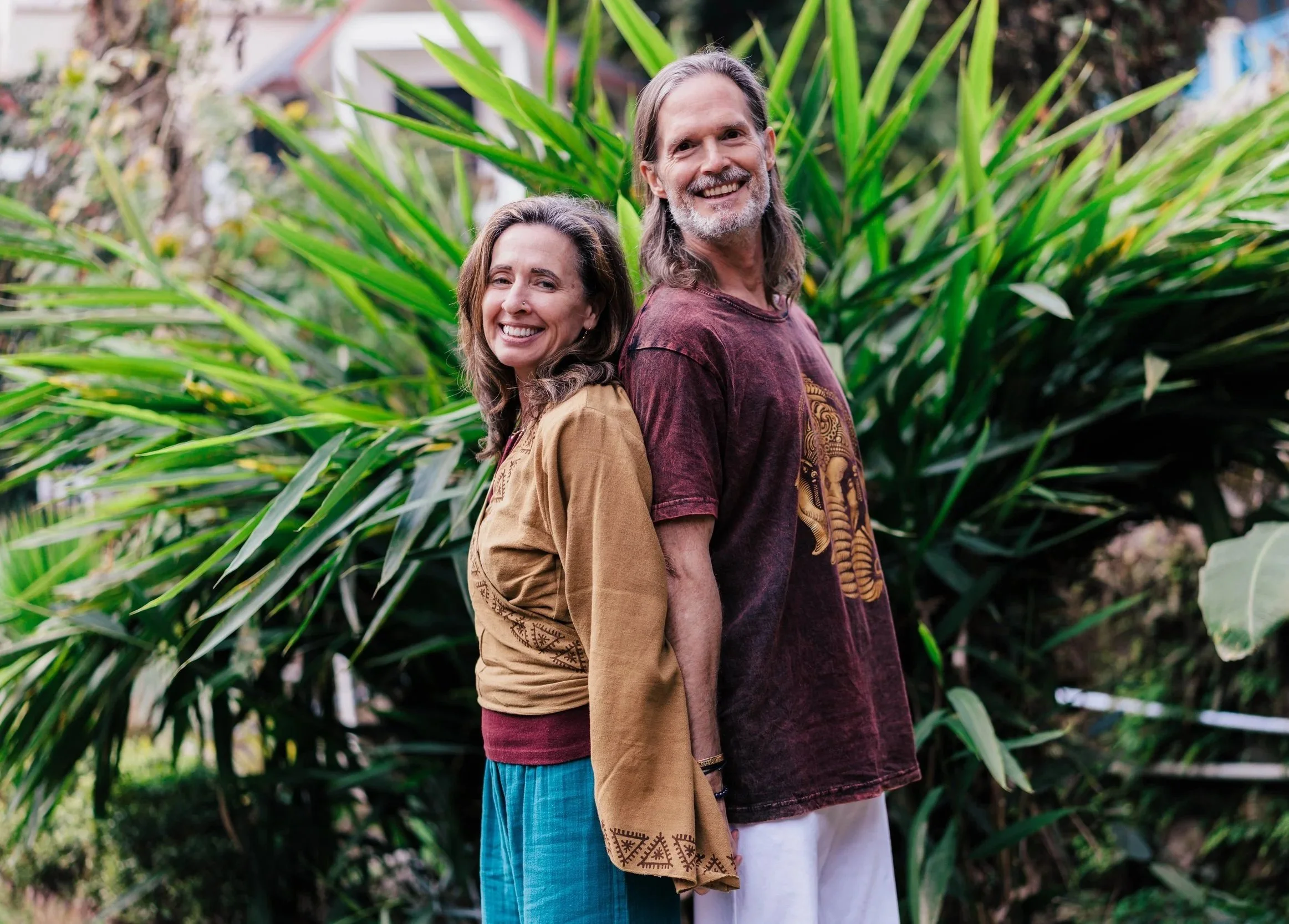 Lexie and Bill standing back-to-back outdoors in front of green tropical plants, dressed in casual, earthy-toned clothing.