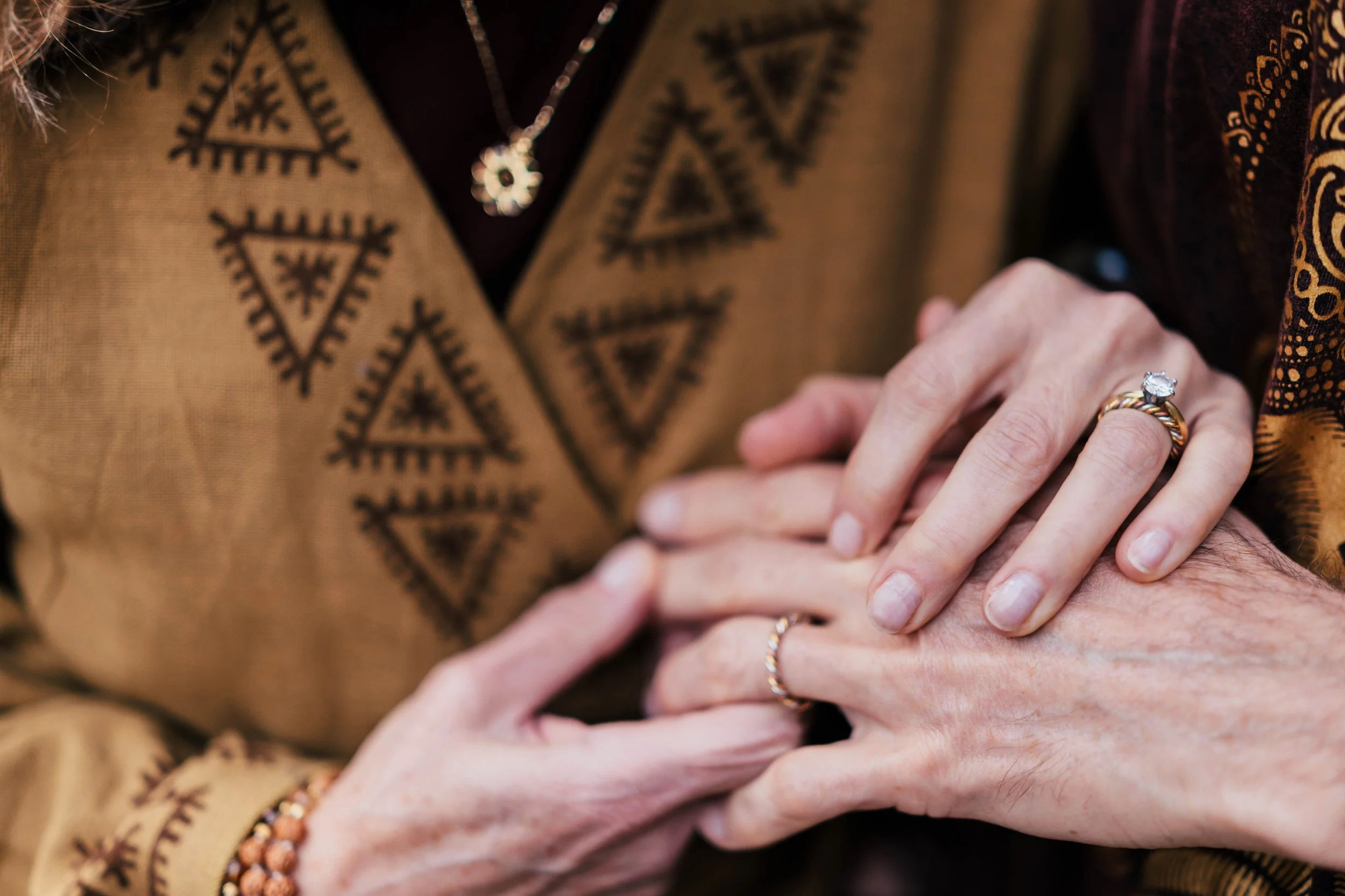 Close-up of multiple hands with rings, intertwined and resting on a person's lap, with patterned clothing in the background.