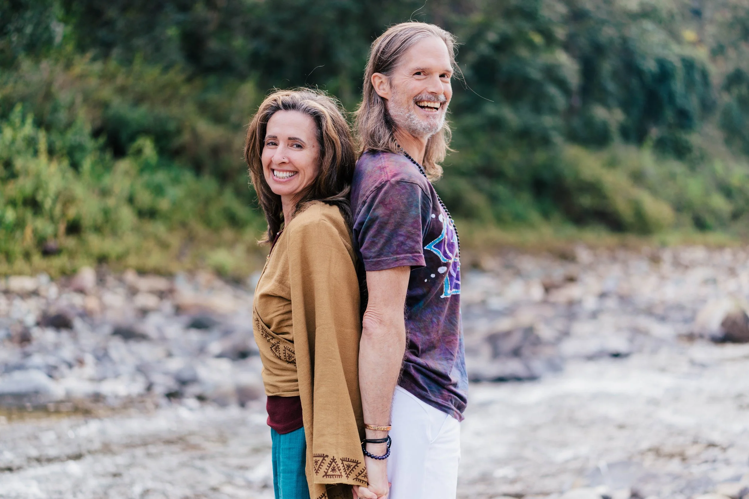 Lexie & Bill standing back to back outdoors on a rocky beach with trees in the background, smiling at the camera.