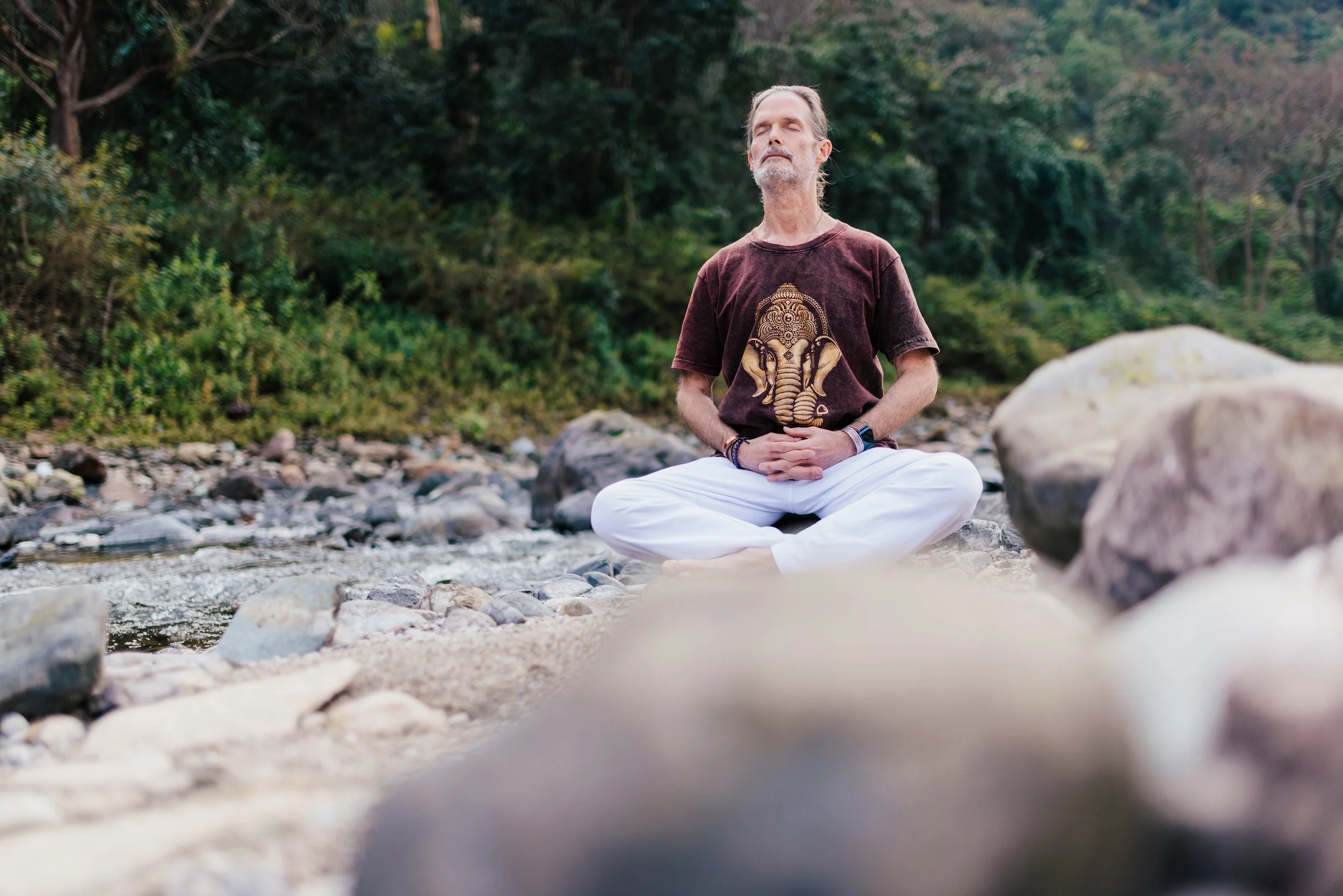 Bill practicing meditation outdoors on a rocky riverbank, surrounded by green trees with his eyes closed and hands resting on his lap.